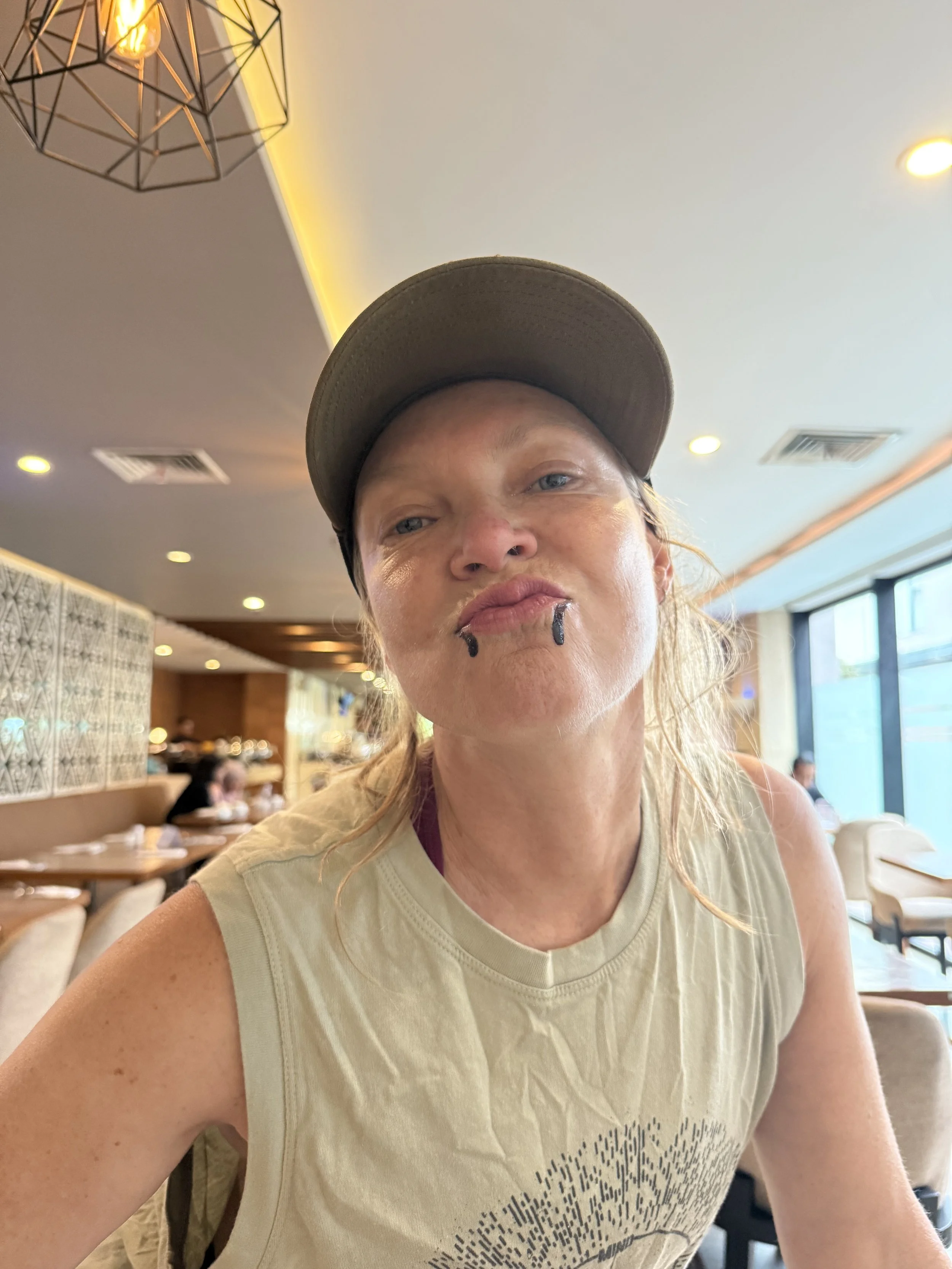 A woman with long blonde hair, wearing a beige sleeveless shirt and a gray baseball cap, is sitting in a restaurant. She has a lip piercing with two black rings and is making a playful, puckered expression. The restaurant has modern decor, large wind