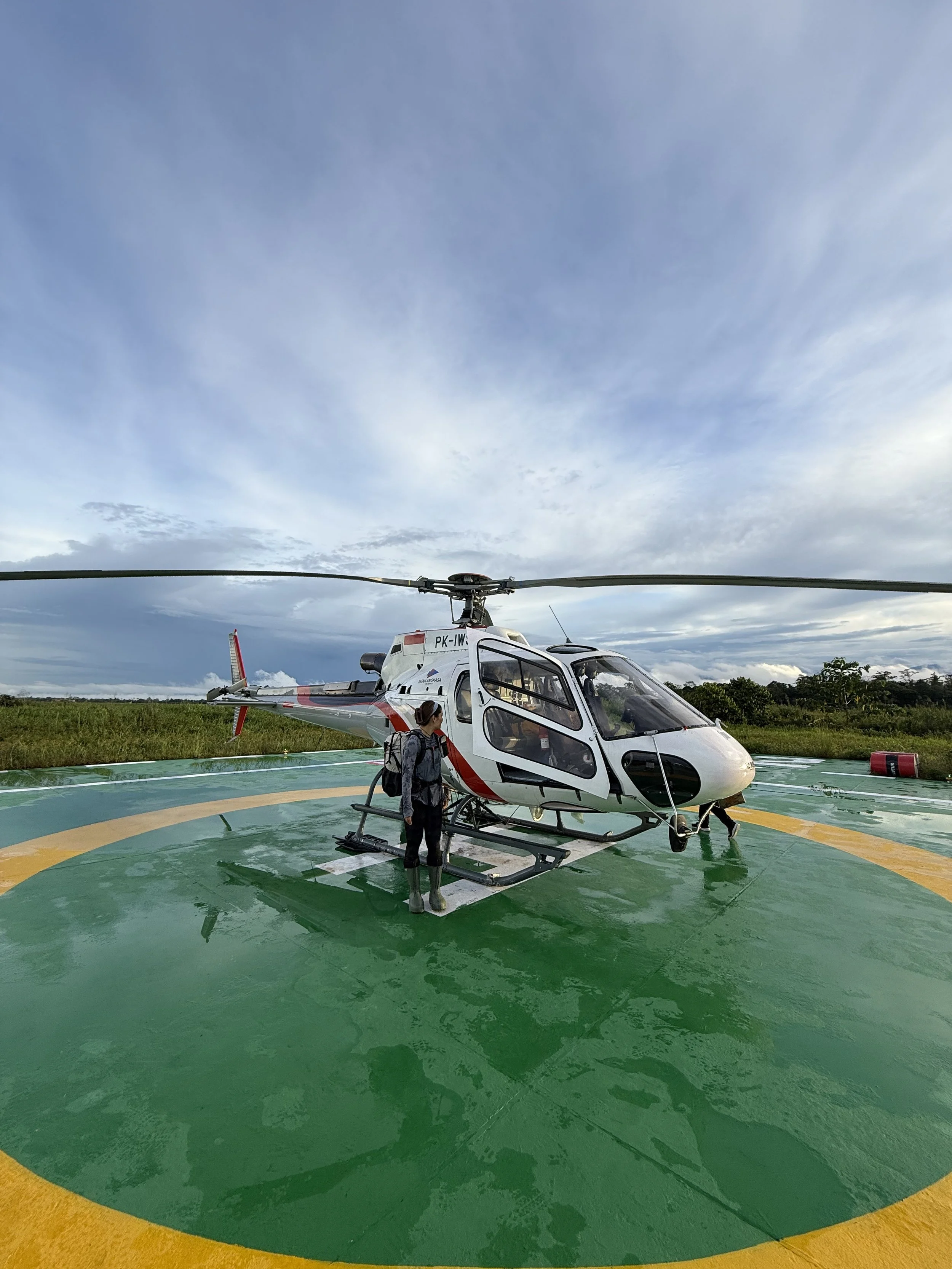 A person standing next to a helicopter on a helipad in an outdoor setting with cloudy sky and greenery in the background.
