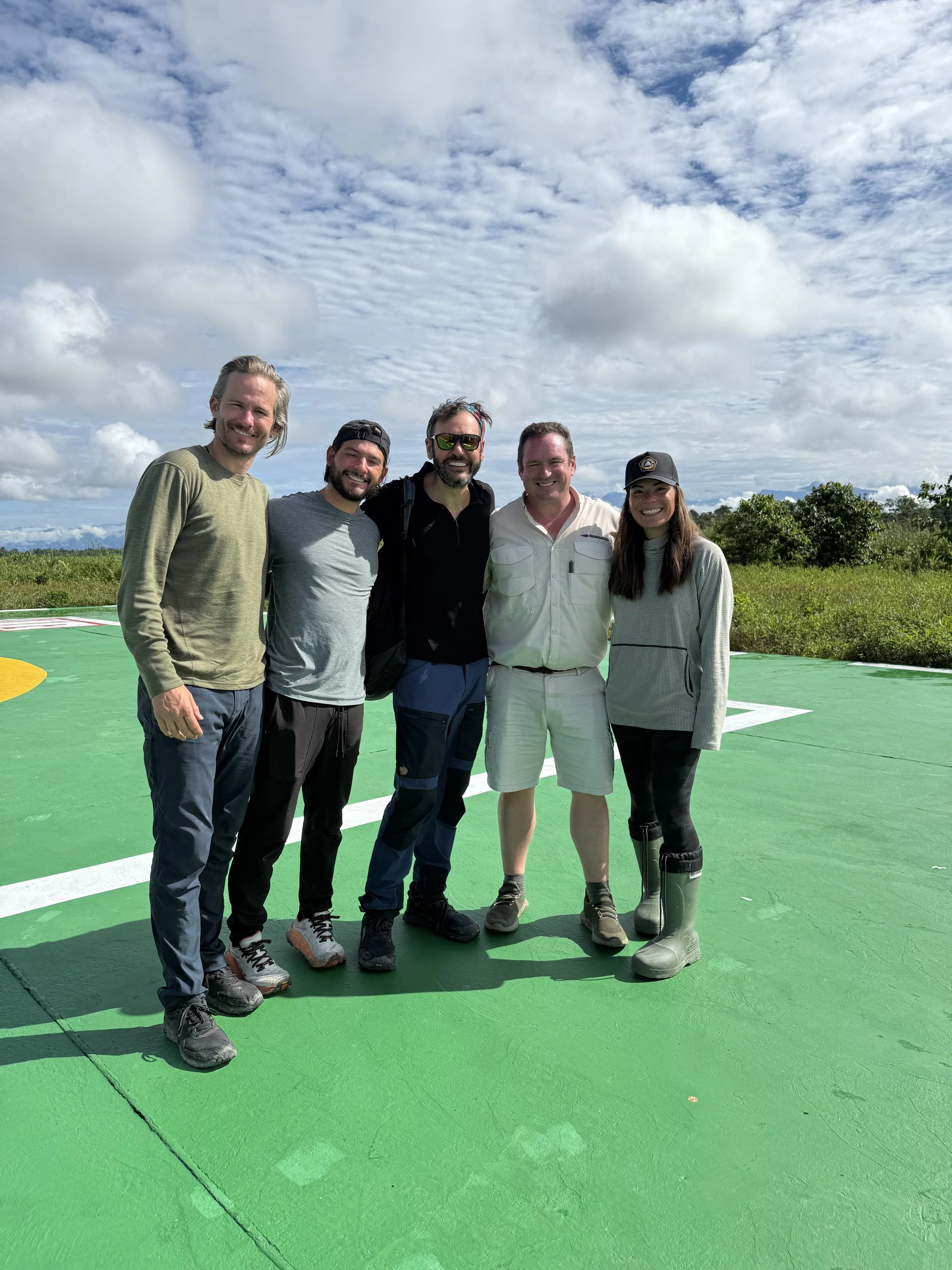 Five people standing outdoors on a green platform with a cloudy sky in the background.