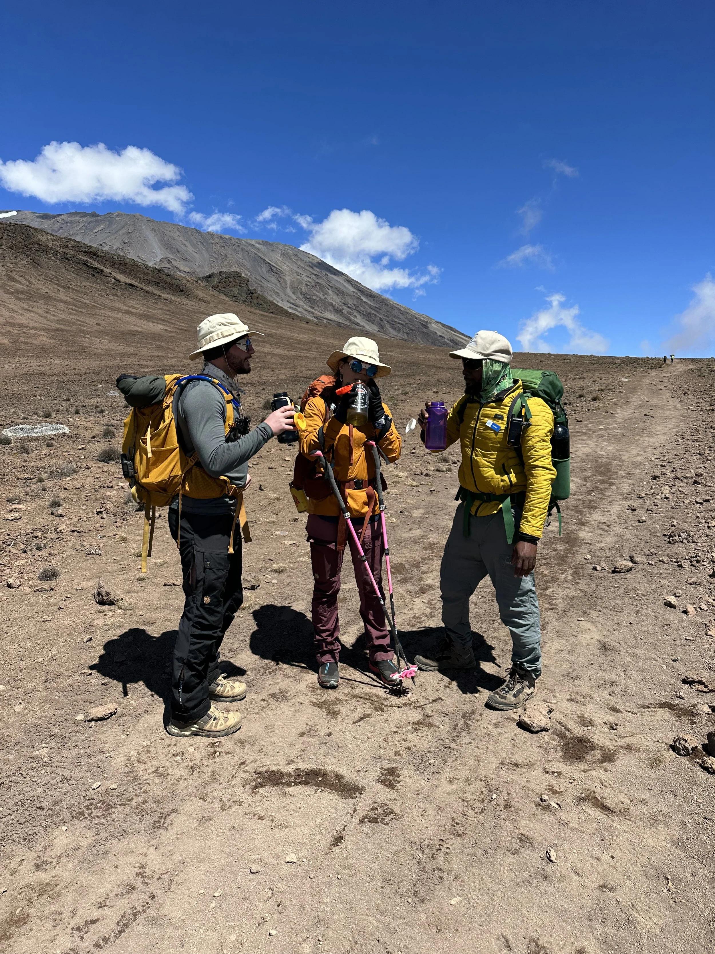 Three hikers in outdoor gear drinking from water bottles in a mountainous, barren landscape under a blue sky.