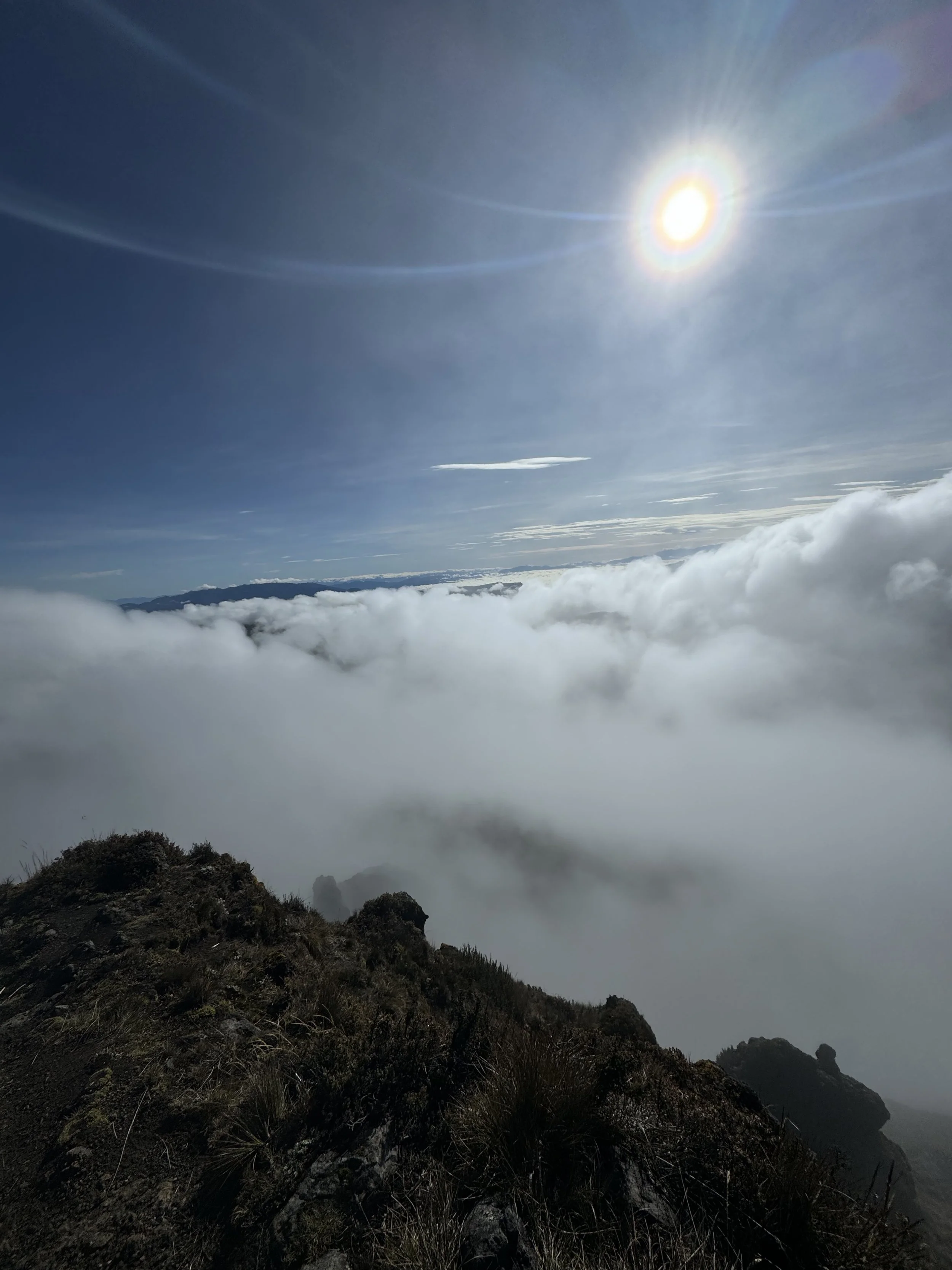 Mountain ridge top with grass and rocks, clouds below, bright sun in clear sky with lens flare.