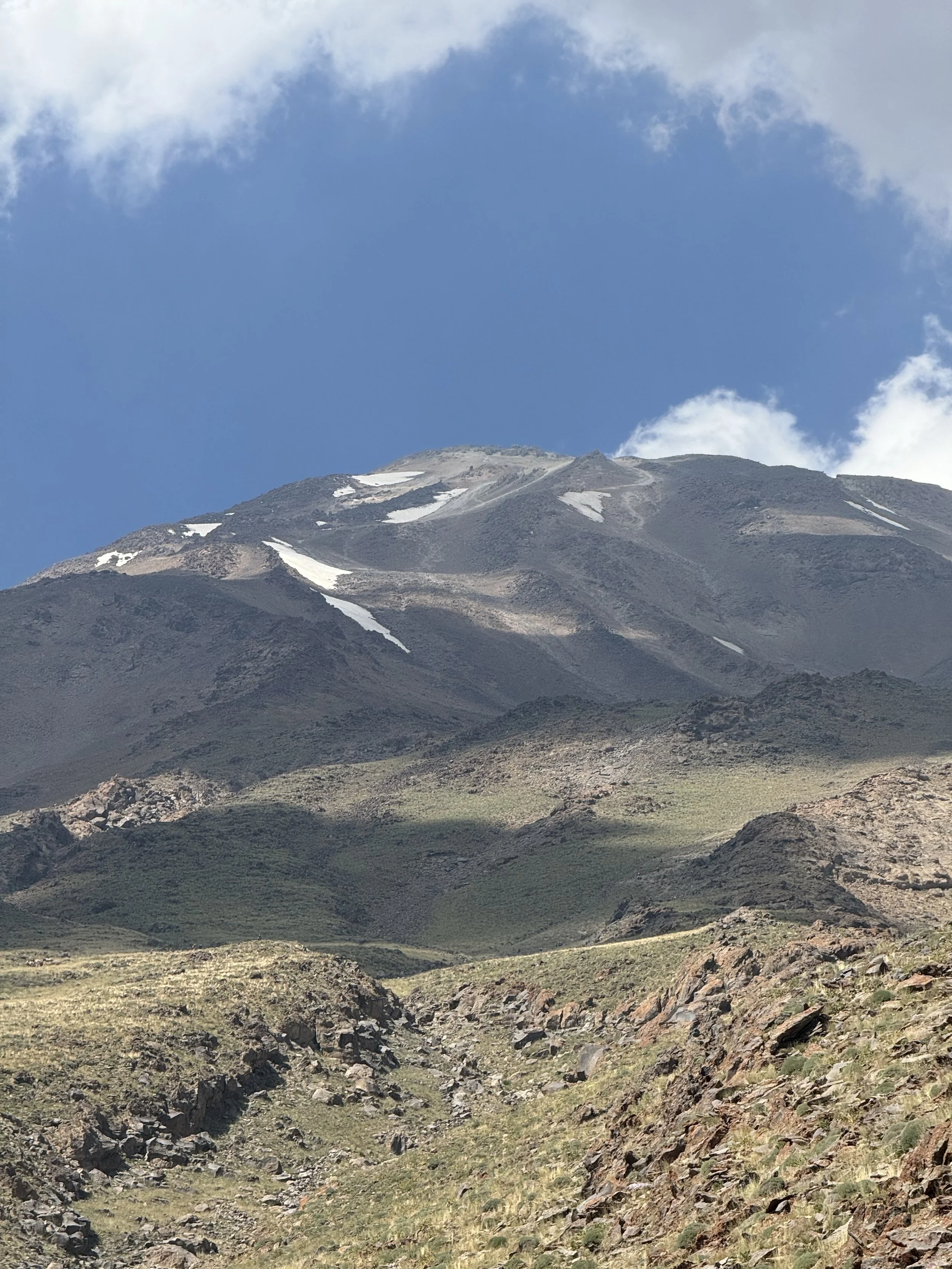 Mountain landscape with a partly snow-capped peak, rocky terrain, and green patches, under a blue sky with some clouds.