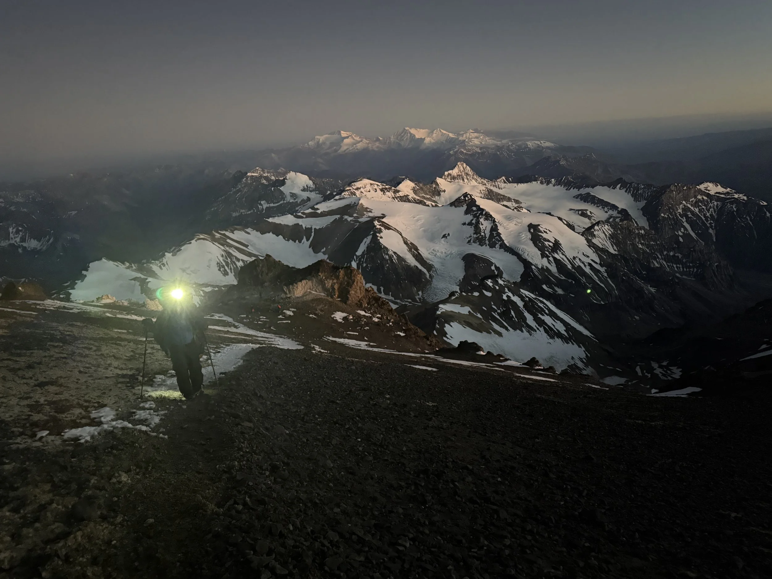 A person with a backpack and hiking poles, wearing a headlamp, trekking on a rugged mountain trail at dusk with snow-capped mountains in the distance.