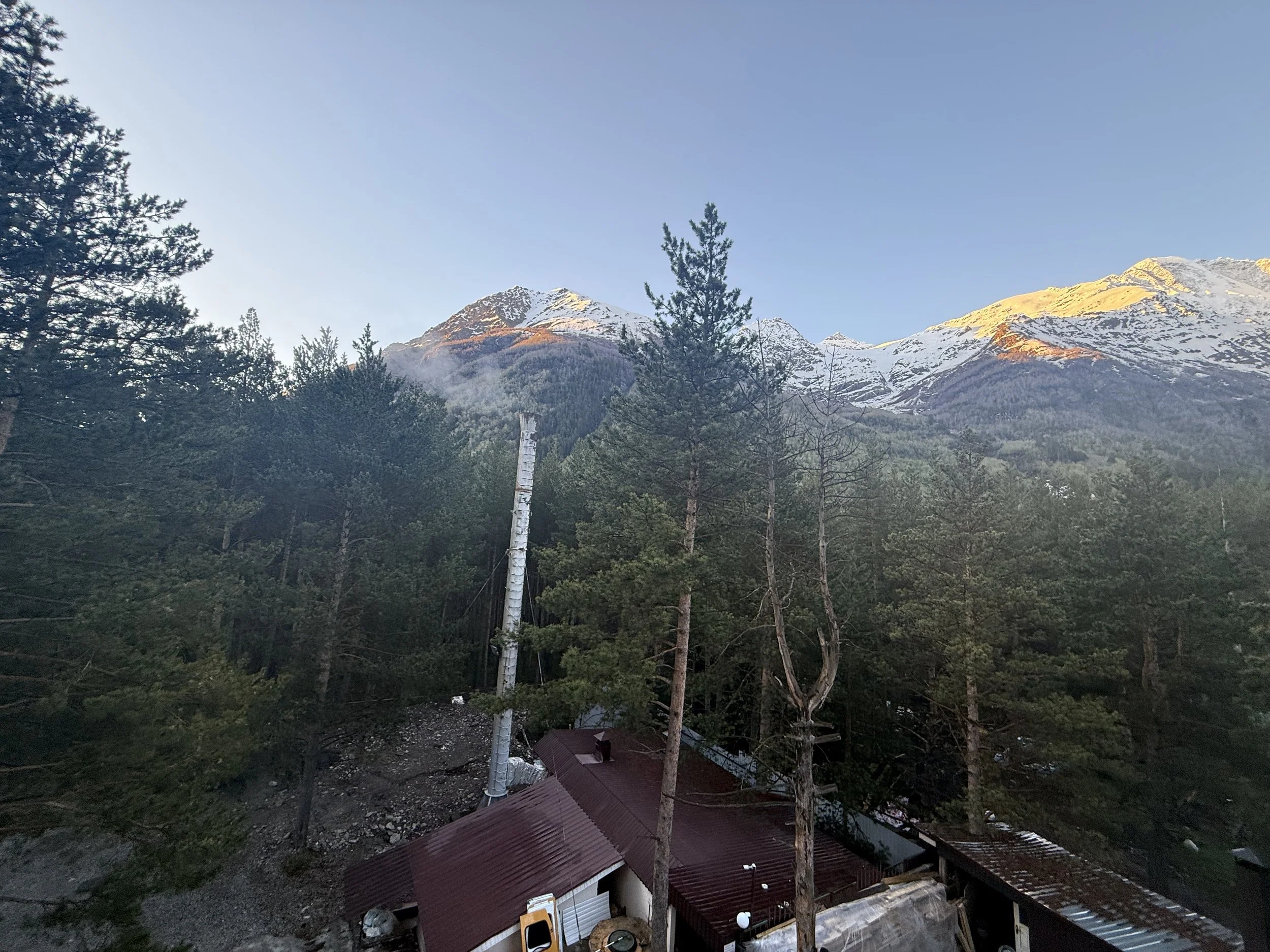 Mountain range with snow-capped peaks in the background, pine trees in the foreground, and a building with a red roof at the bottom.