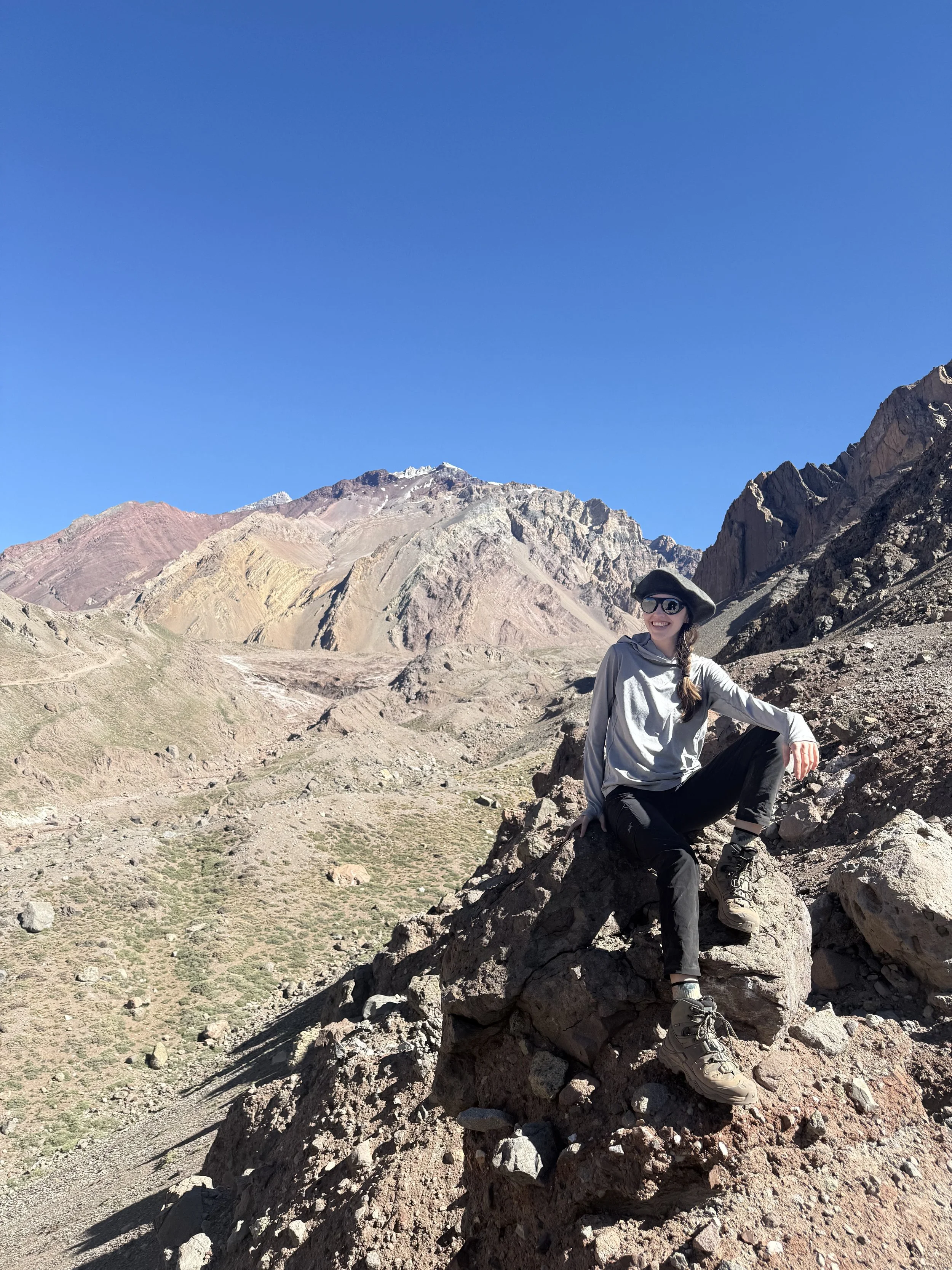 A woman sitting on a large rock in a mountainous desert landscape with colorful mountain range and clear blue sky behind her.