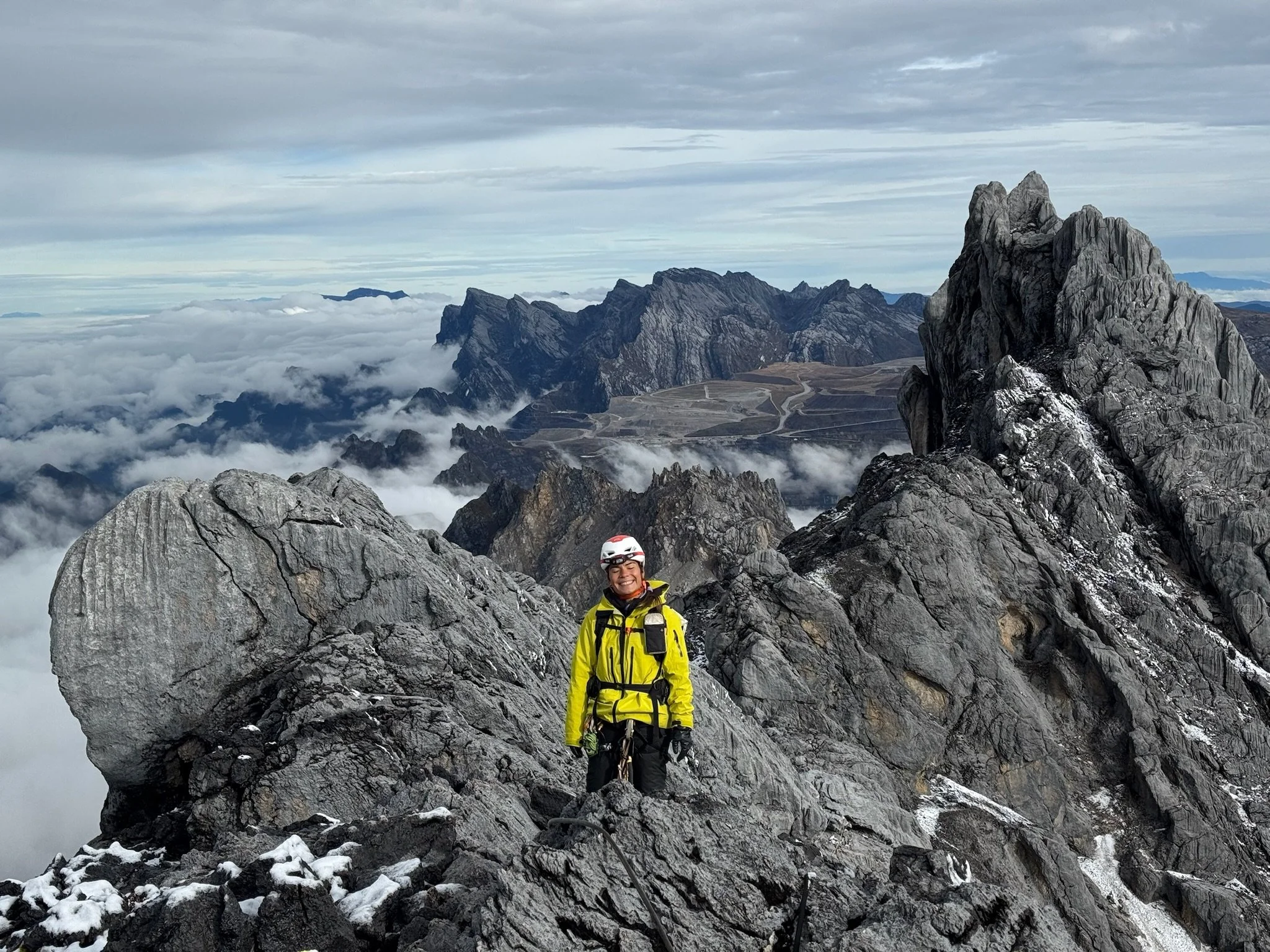 A person wearing a yellow jacket and helmet standing on a rocky mountain peak with a view of other mountain ridges and clouds in the background.
