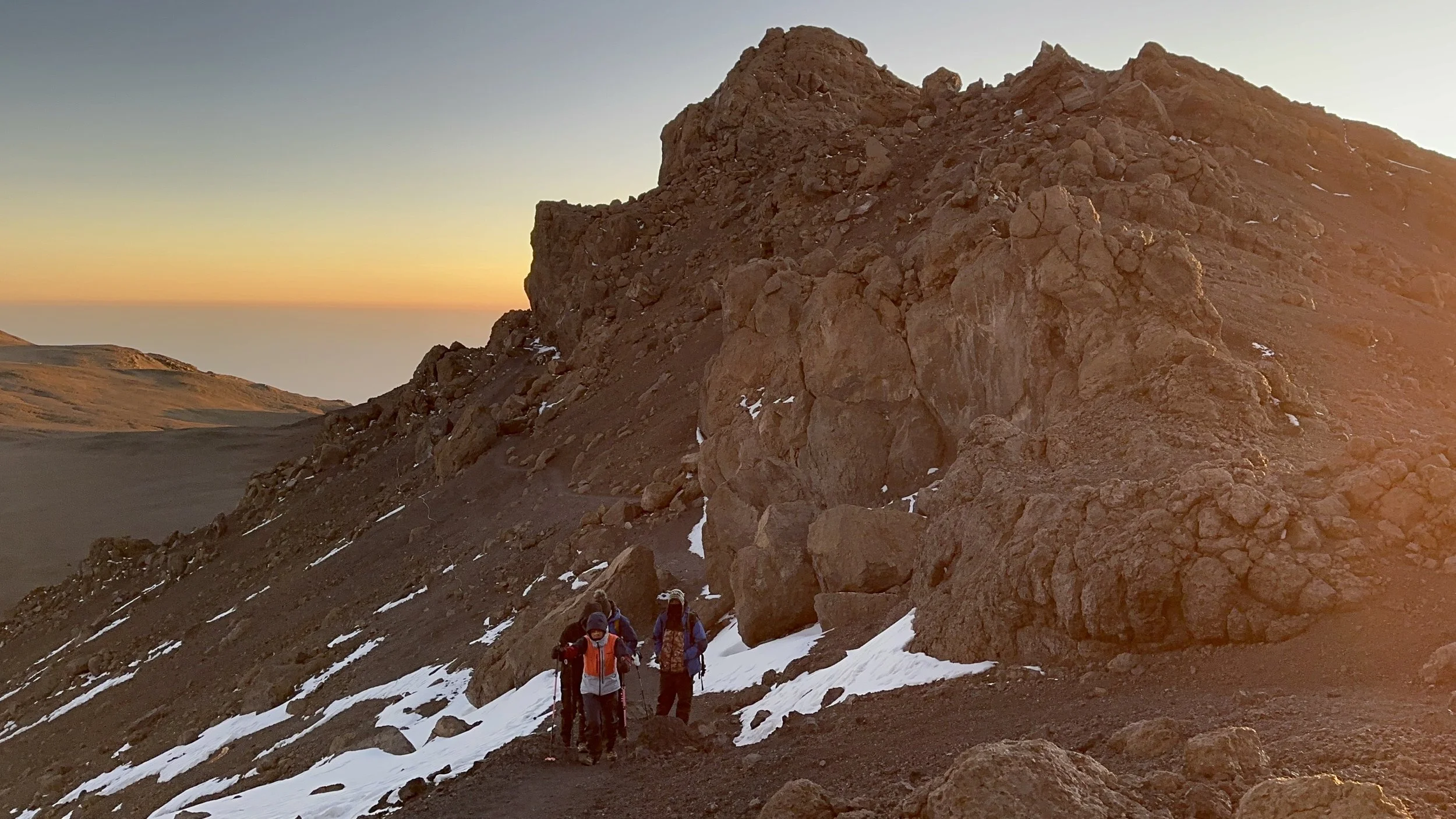Group of four hikers walking on a mountain trail with snow patches, large rocks, and a sunset sky in the background.