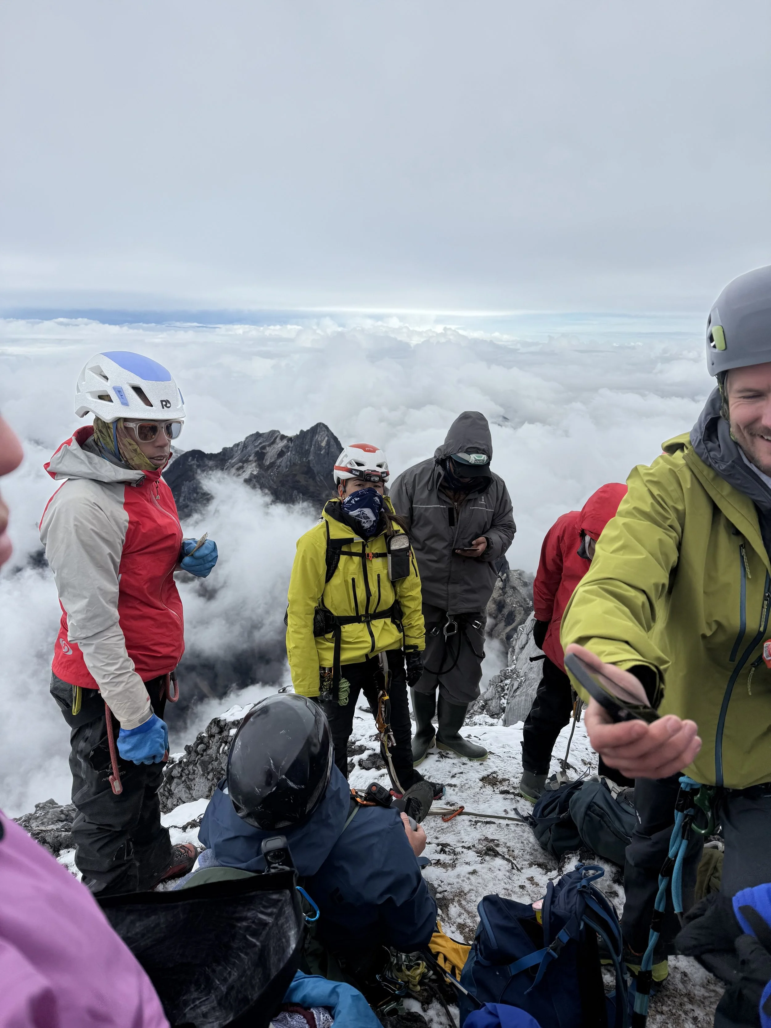 Mountain climbers in gear resting on snow-covered peak with clouds and distant mountain peaks in the background.