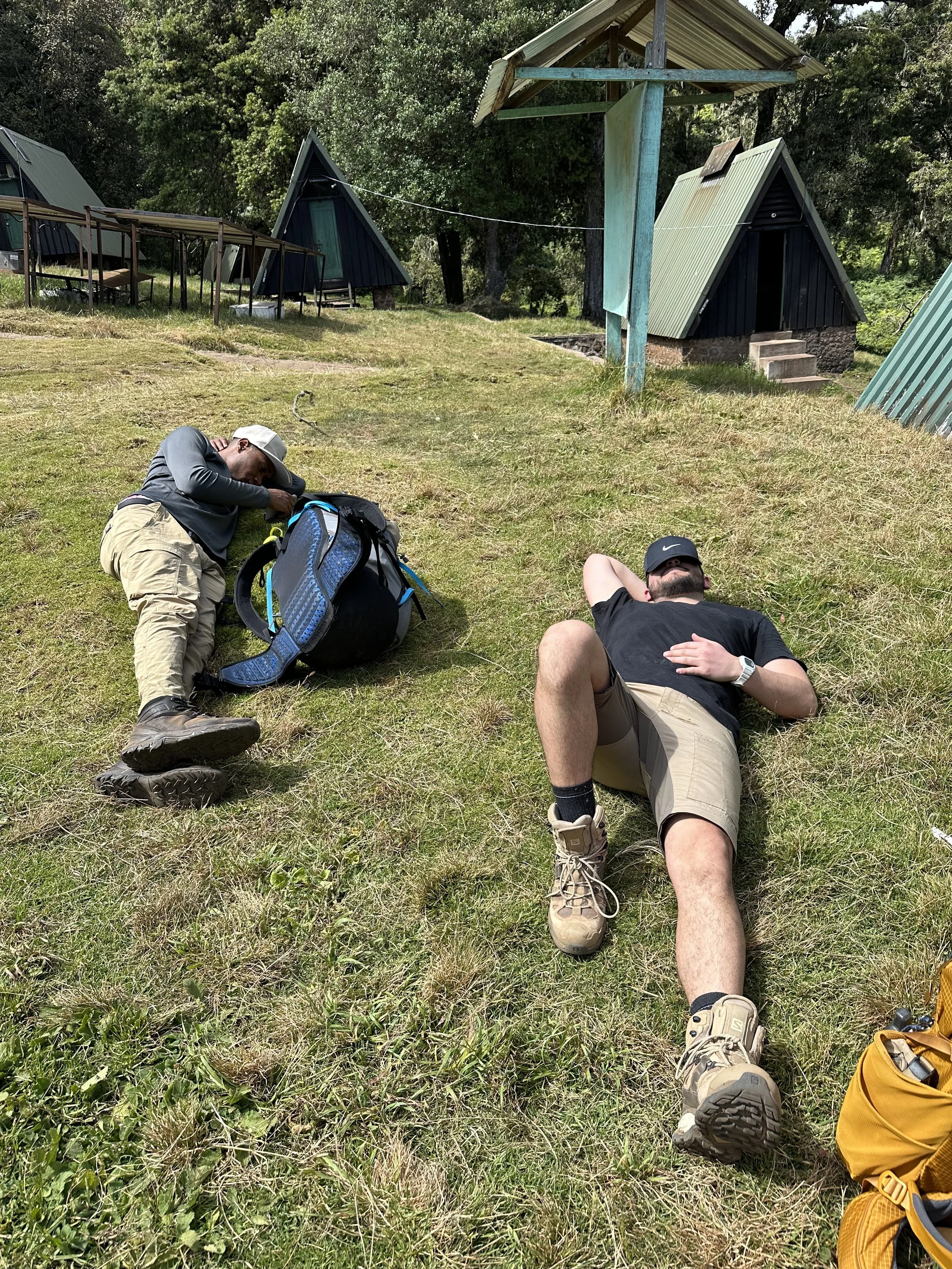 Two men lying on the grassy ground near small cabin-like structures in a forested area, resting after outdoor activities.