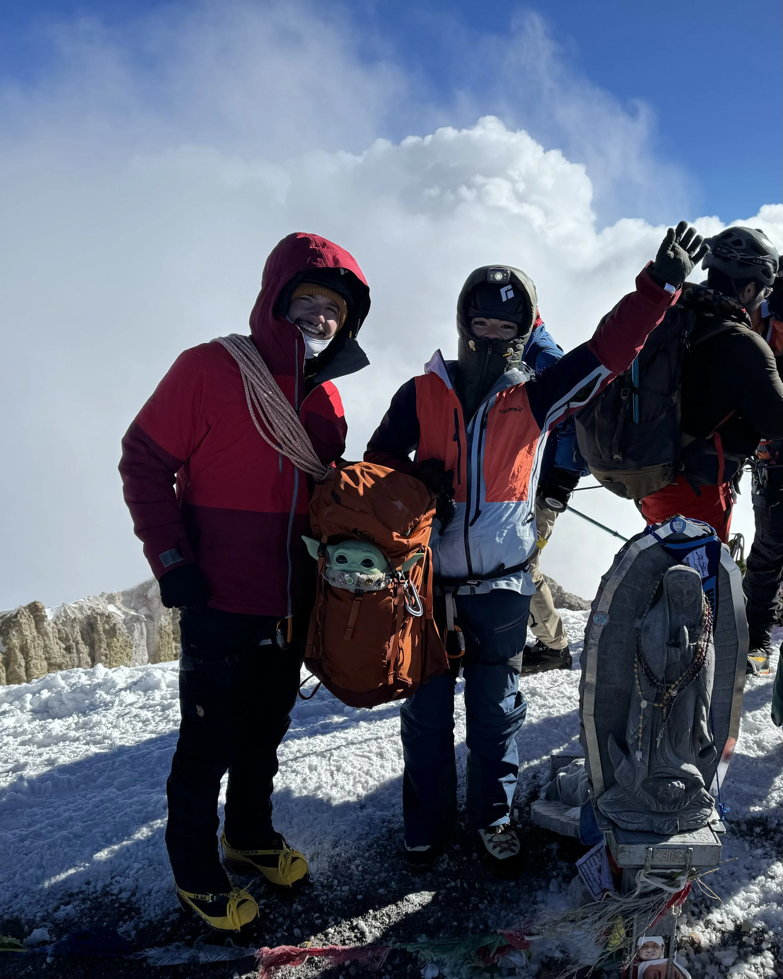 Two climbers standing on a snow-covered mountain summit, wearing colorful insulated jackets, snow boots, and helmets. One holds an orange backpack with a helmet attached, and another is making a peace sign with their glove. A cloudy sky is visible be