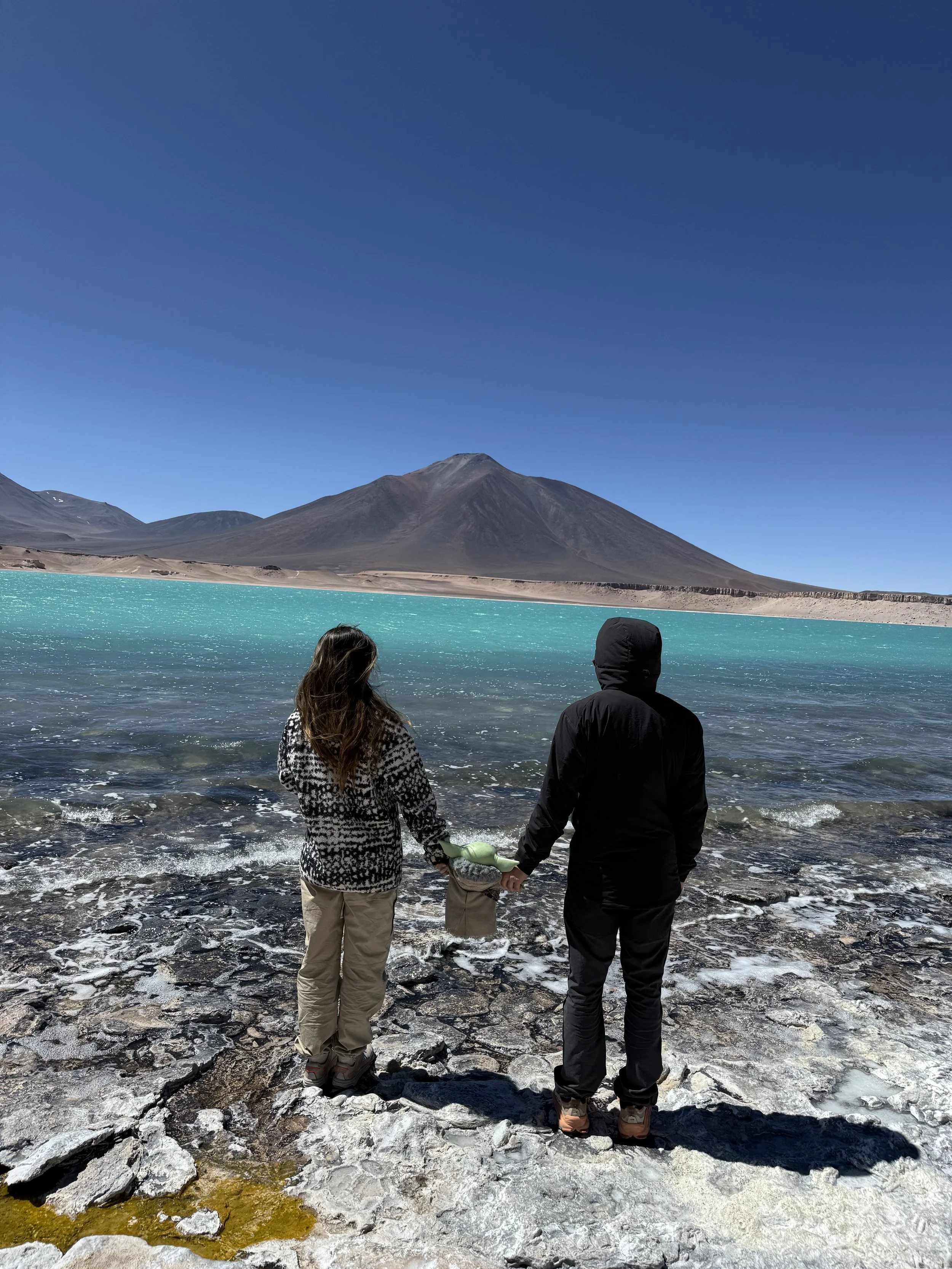 Two people, a woman and a man, holding hands at the edge of a turquoise lake with volcanic mountains in the background.