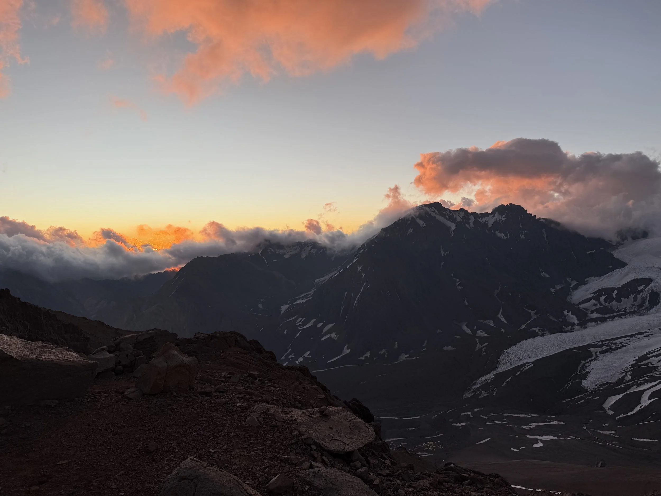 Sunset over rugged mountain landscape with rocky foreground, snow patches, and clouds.