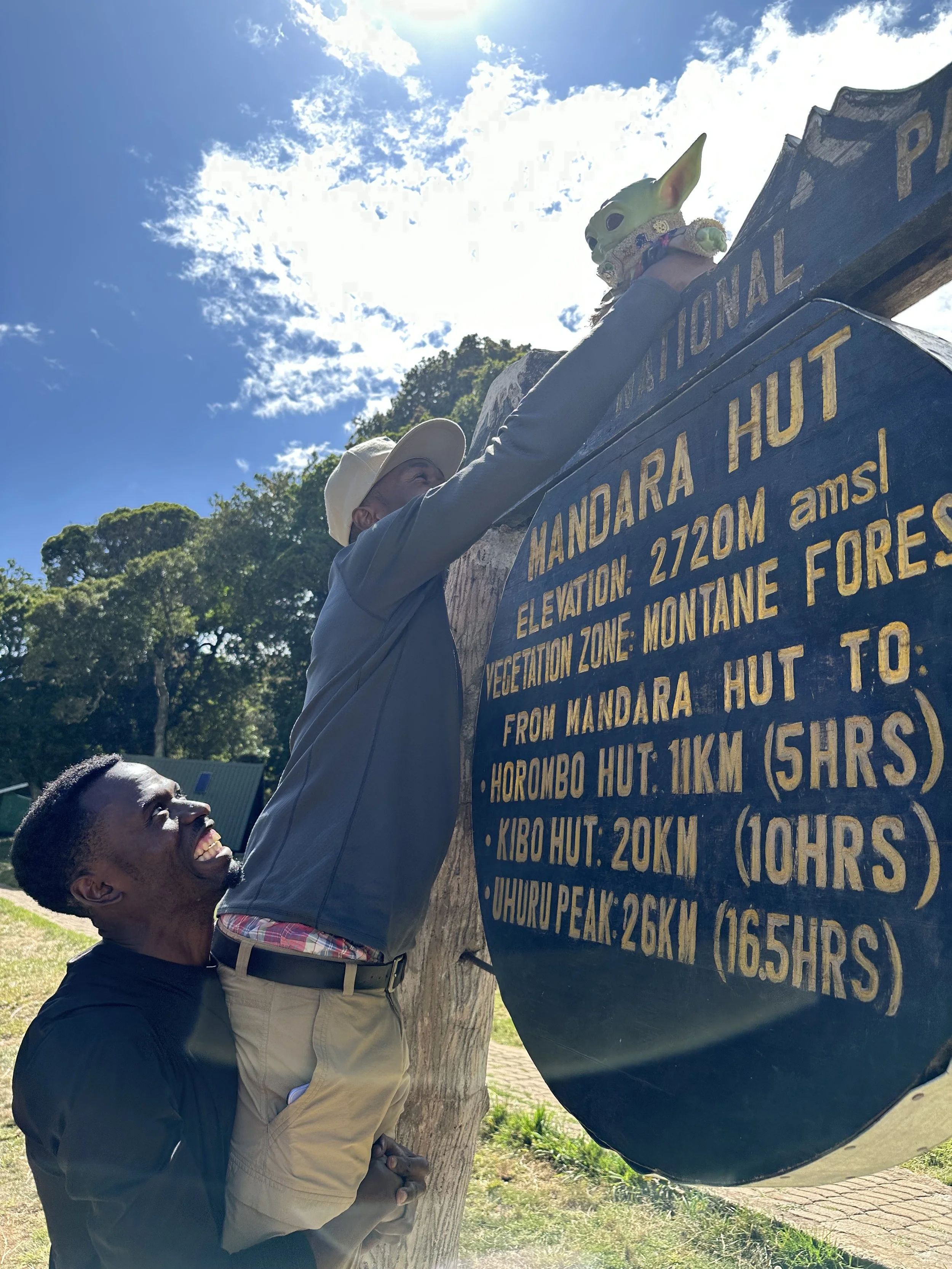 Two men, one standing on the other's shoulders, are hanging a figurine of a creature with large ears and a mask on a signpost at Mandara Hut. The signpost provides information about the elevation and hiking distances to various huts at Mount Kilimanj