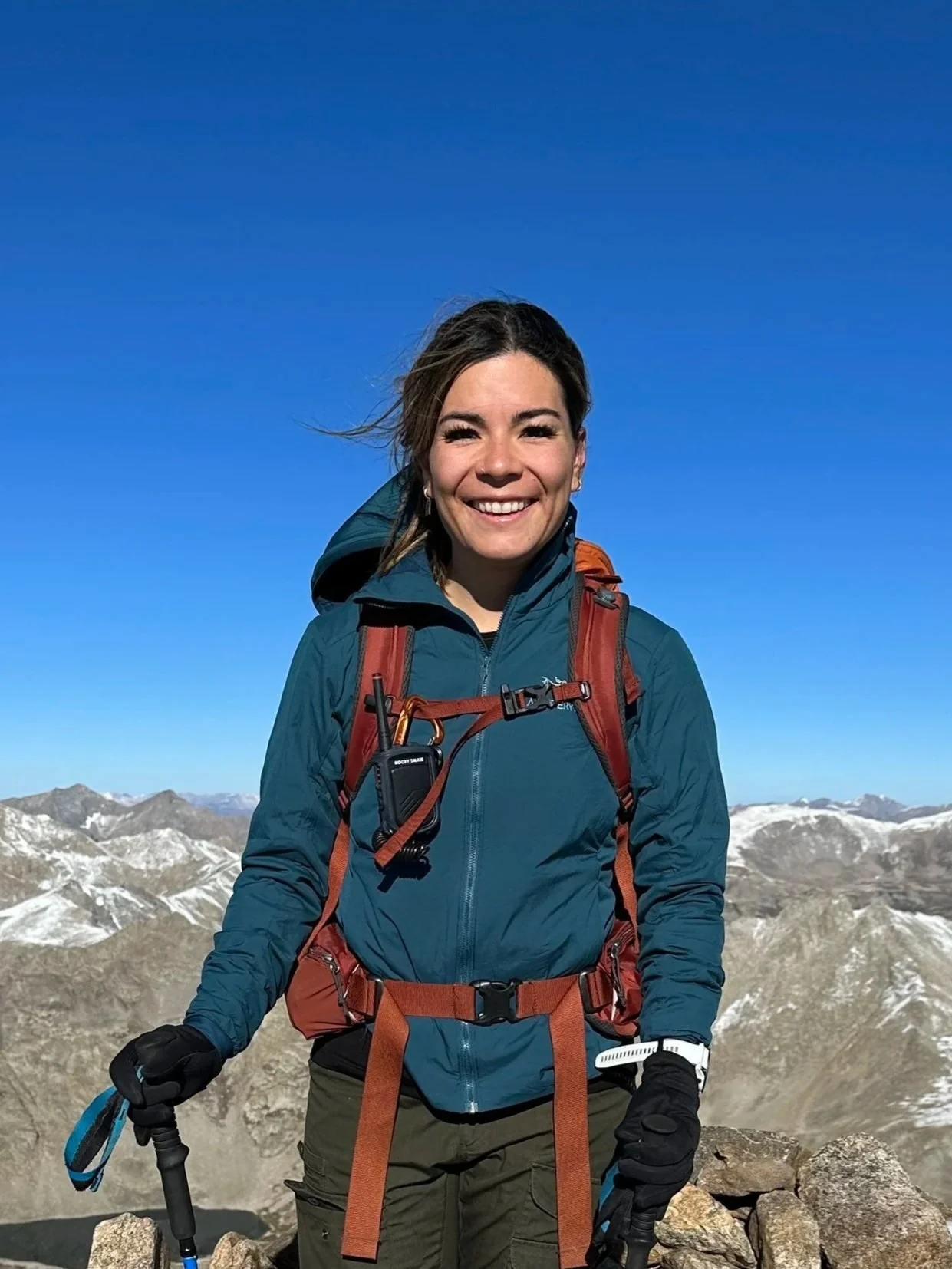 A woman in outdoor hiking gear standing on rocky mountain summit with snow-capped peaks in the background under a clear blue sky.