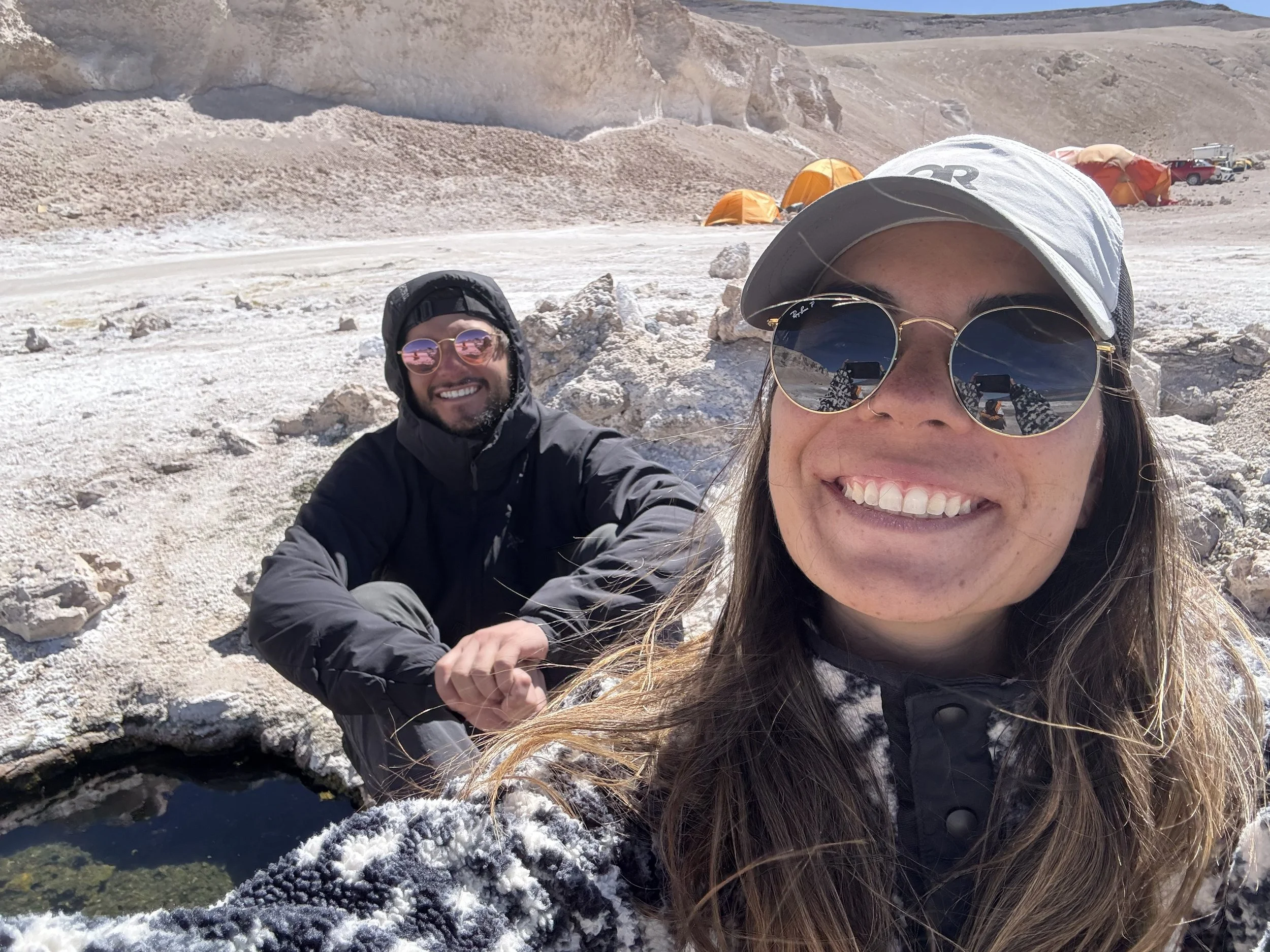Two people posing on a snowy landscape with tents and rocky formations in the background, smiling and wearing outdoor gear and sunglasses.