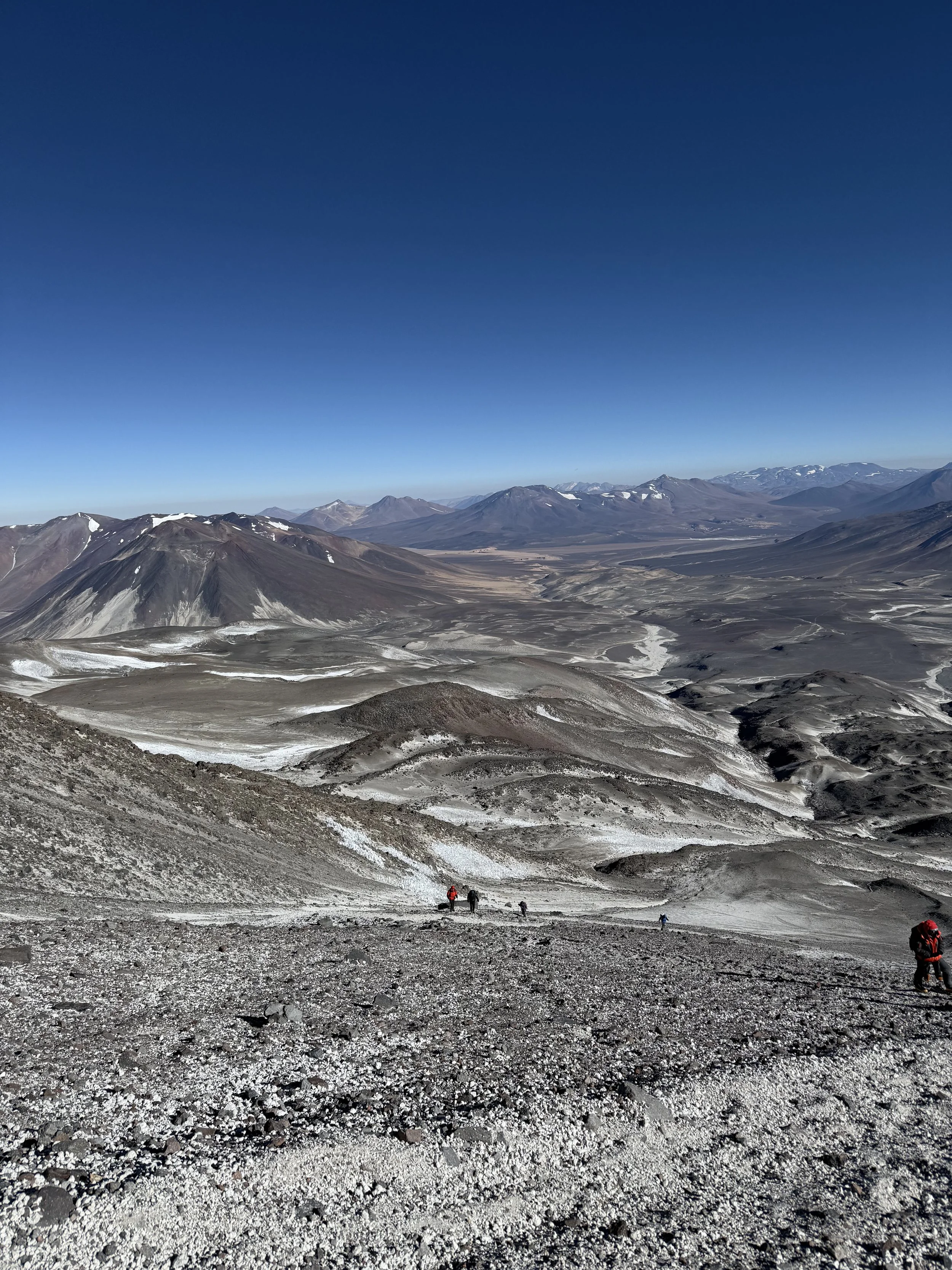 Mountains with a dry, rocky foreground and a clear blue sky, with hikers climbing a slope.