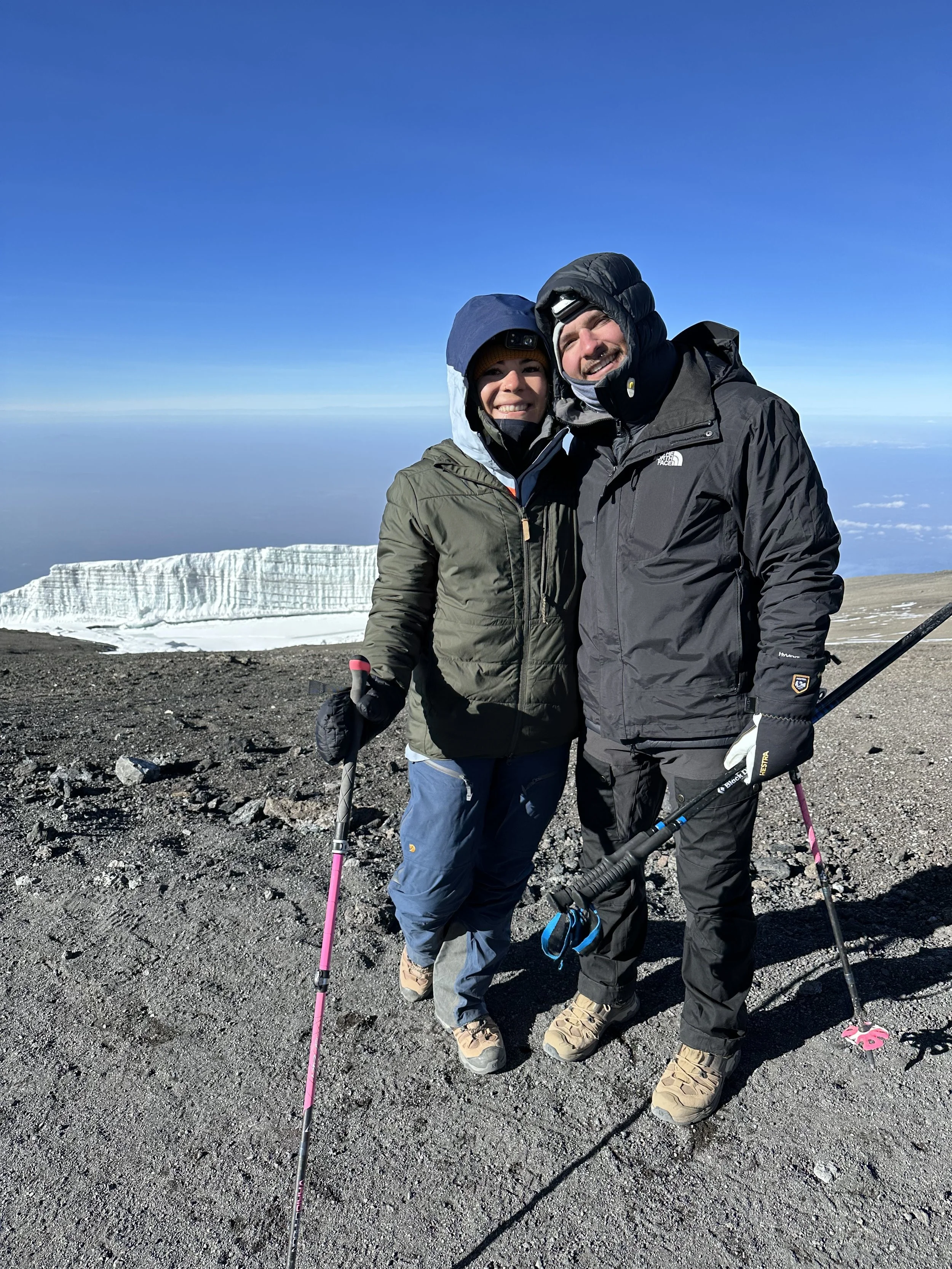 Two hikers in winter gear standing on a mountain summit with snow and ice in the background under a clear blue sky.