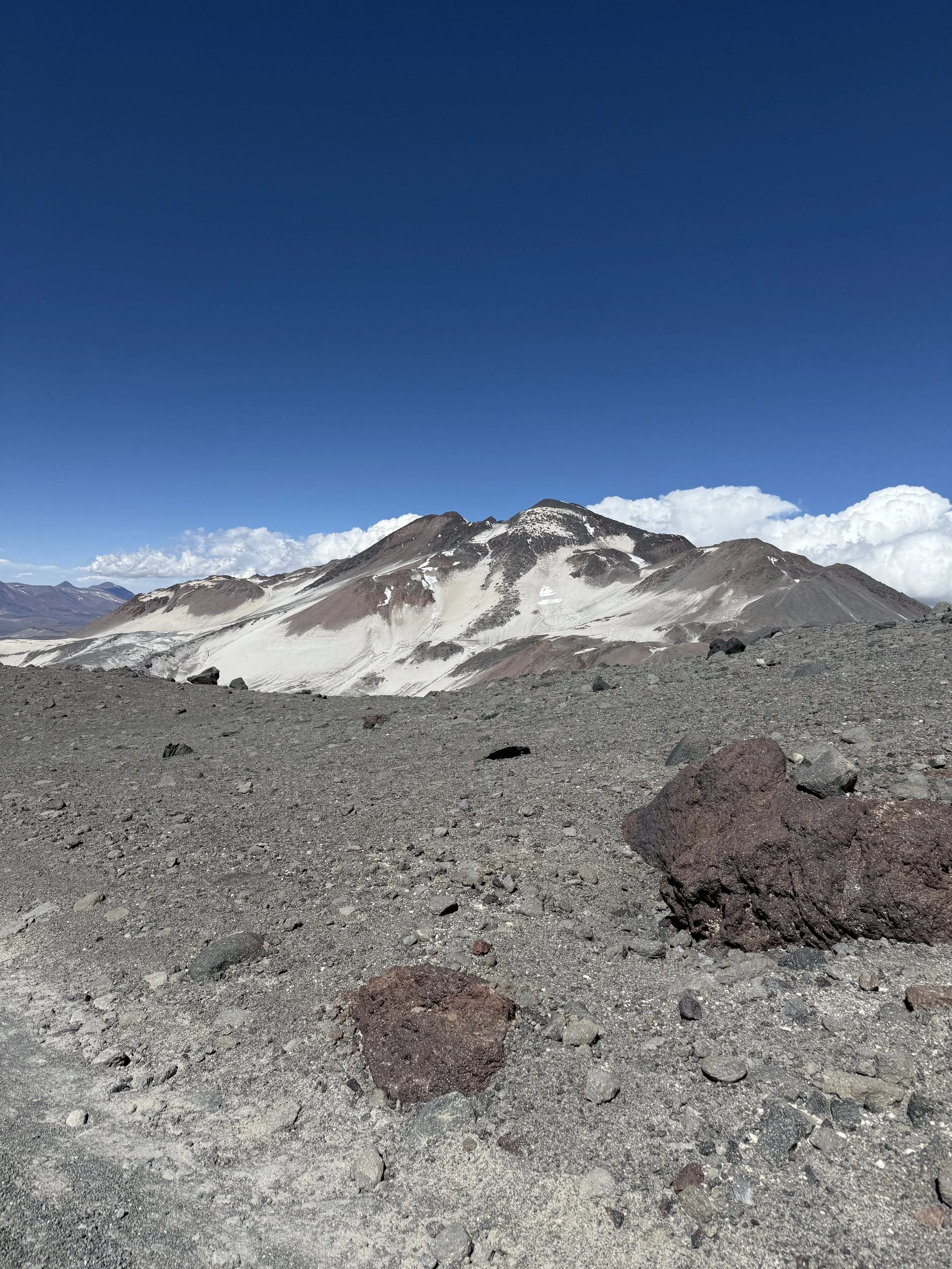 A barren, rocky desert landscape with large rocks in the foreground and a mountain range with patches of snow in the background under a clear blue sky.