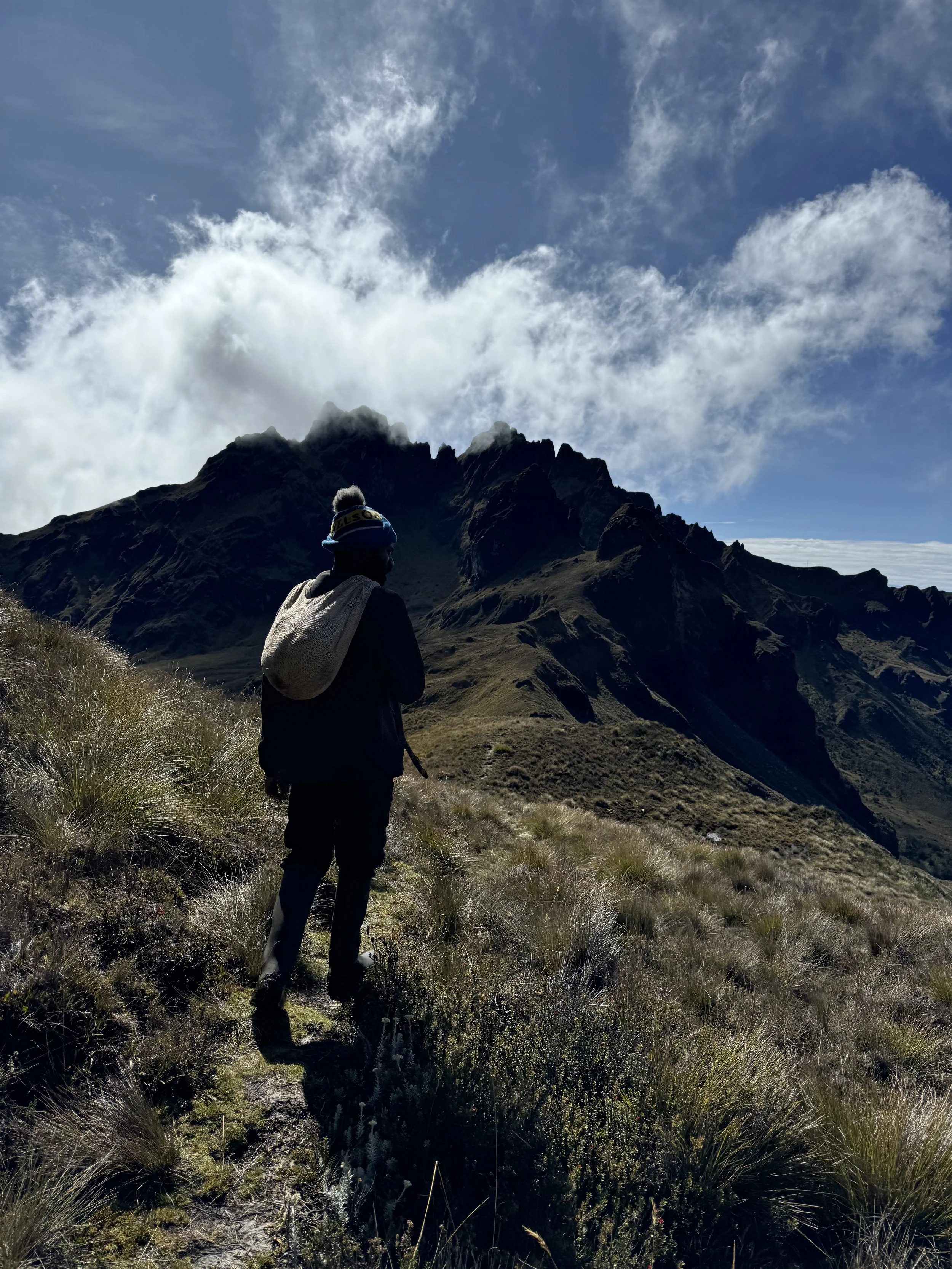 A person hiking on a mountain trail with rugged mountains and a partly cloudy sky in the background.