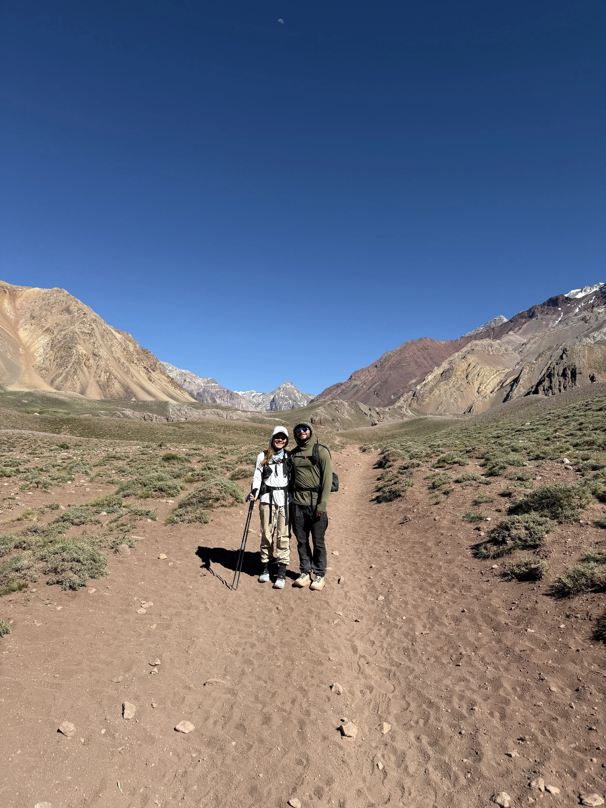 Two hikers standing on a dirt trail in a mountainous desert landscape under a clear blue sky.