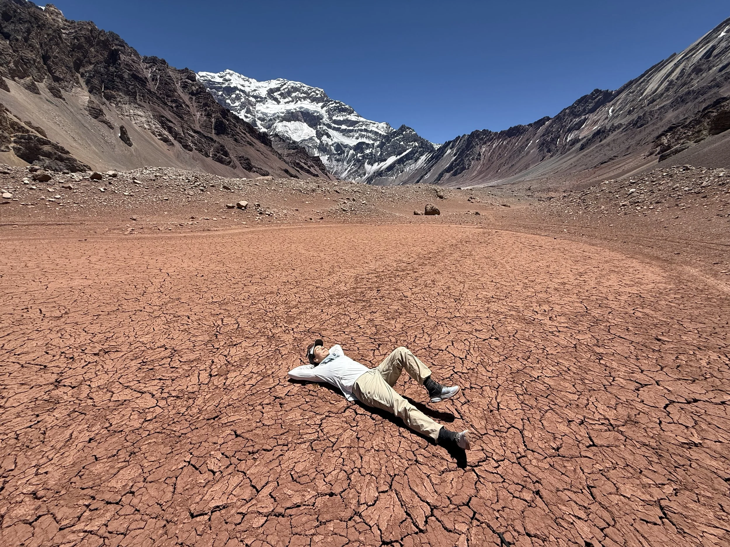 Person lying on cracked, reddish ground in a mountainous desert with snow-capped peaks under a clear blue sky.