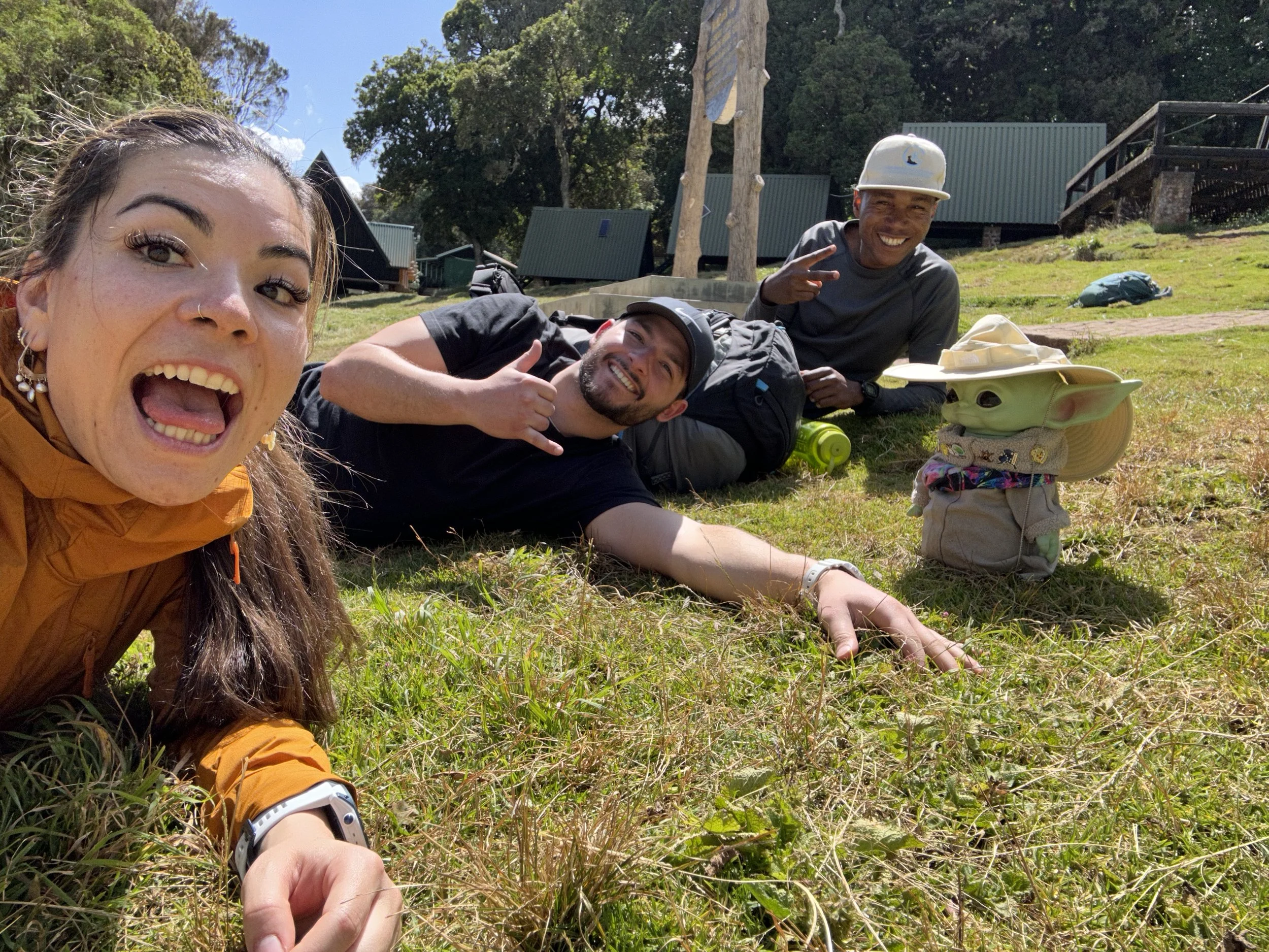 Three friends lying on the grass outdoors, smiling and making peace signs, with tents and trees in the background at a campsite on a sunny day.
