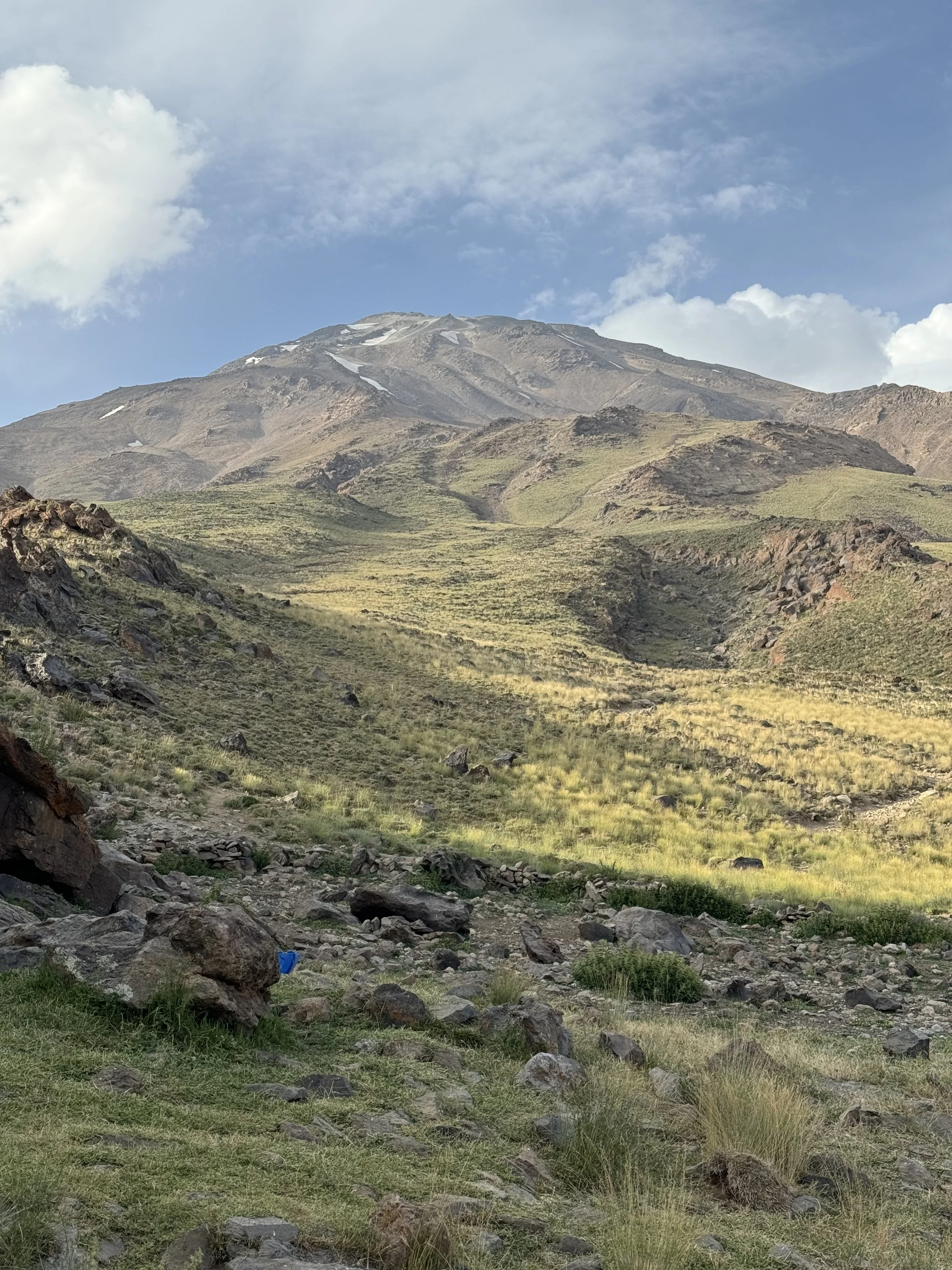 A mountainous landscape with a large mountain in the background, grassy slopes, rocks in the foreground, and a partly cloudy sky.