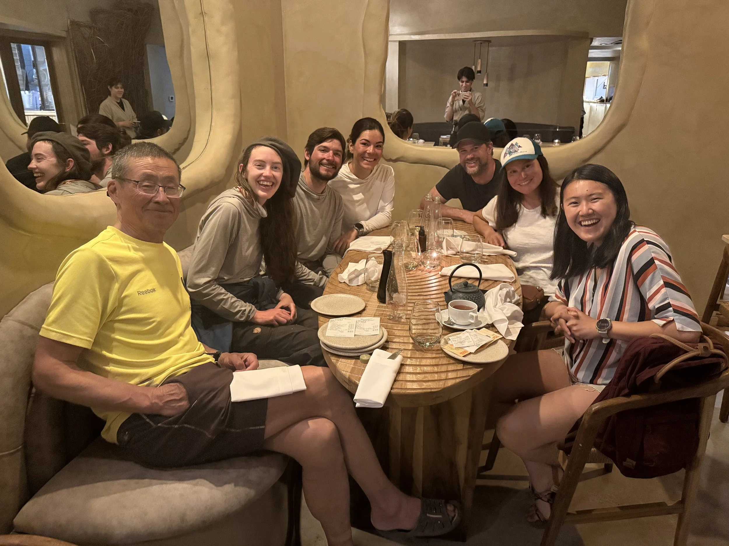 A group of nine people sitting around a wooden table at a restaurant, smiling and enjoying the meal. The table is set with glasses, cups, and plates, and there are two mirrors on the wall behind them reflecting some of the other guests and restaurant