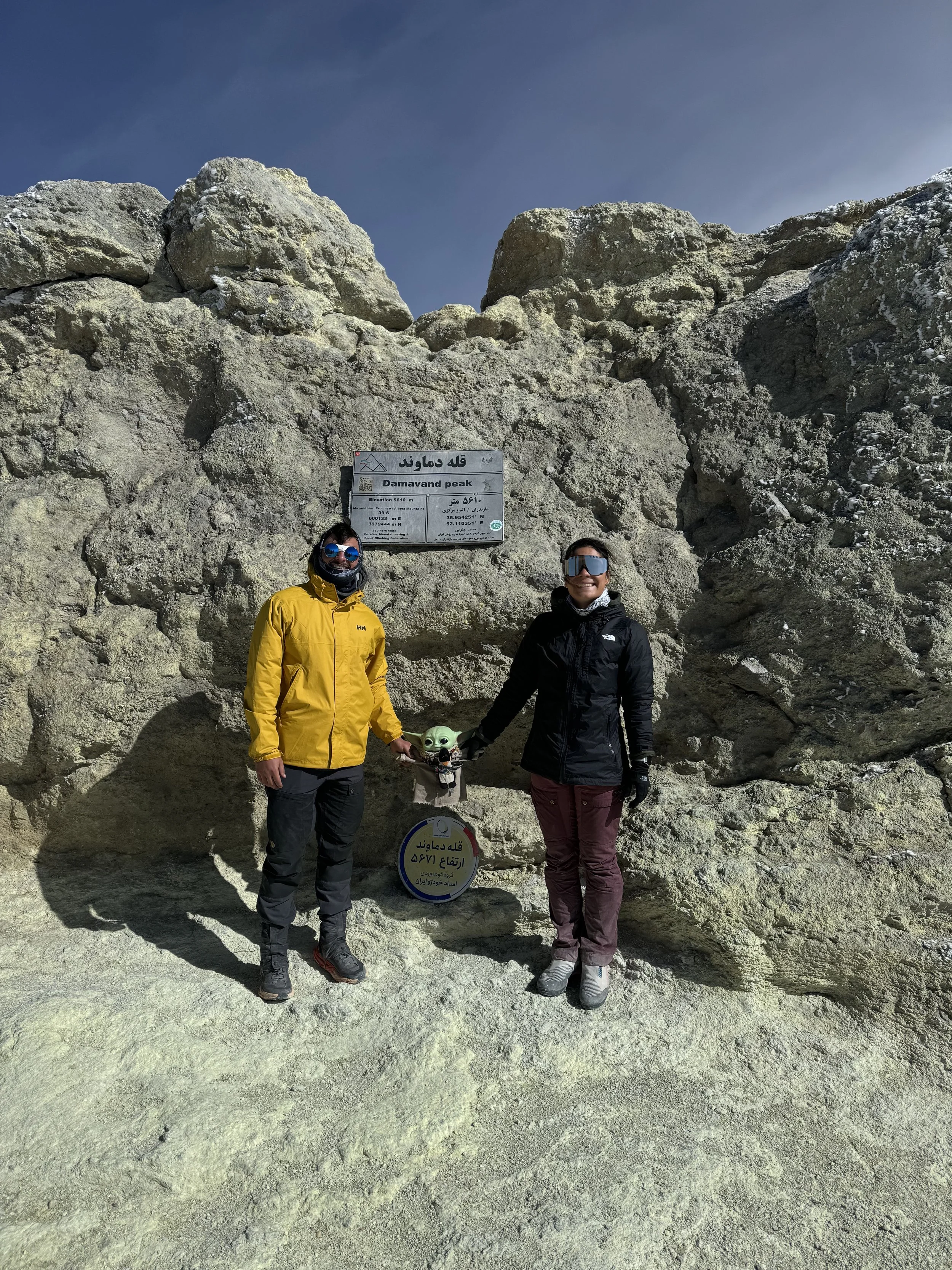 Two people standing at Damavand Peak, holding a small green Yoda-shaped sculpture, with rocky terrain and a sign indicating the mountain's elevation in the background.