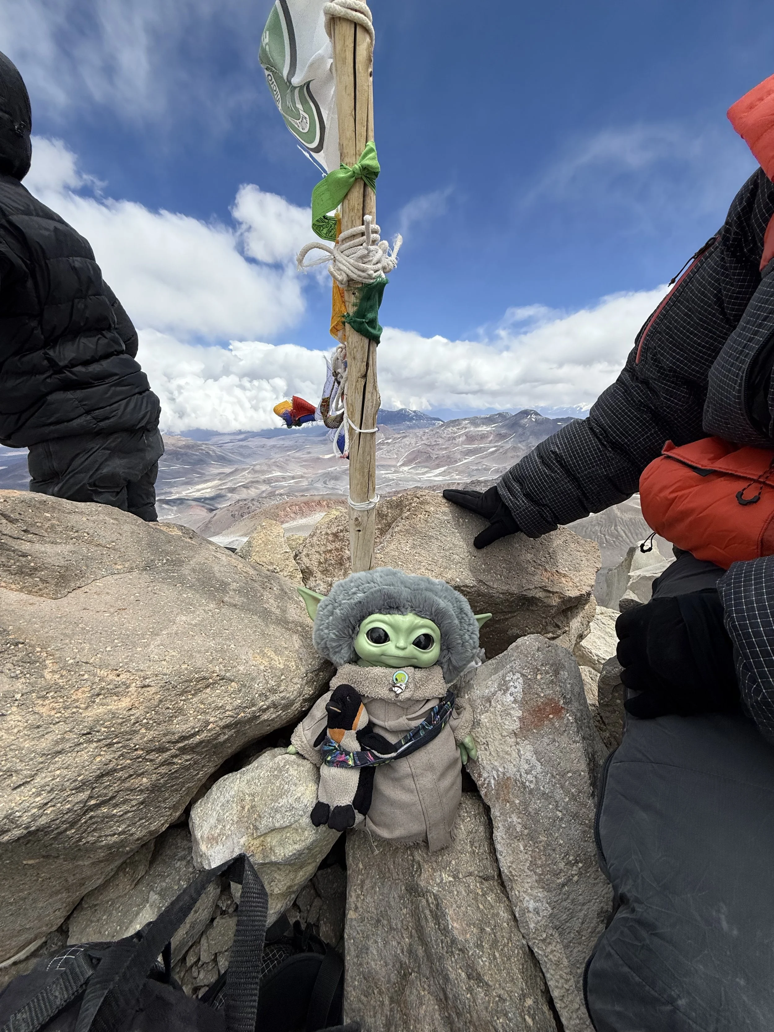 A plush toy of Baby Yoda (Grogu) from Star Wars, dressed in a cozy outfit with a scarf, sitting among rocks on a mountain summit. There are two hikers in winter gear on either side, with a mountain landscape and cloudy sky in the background.