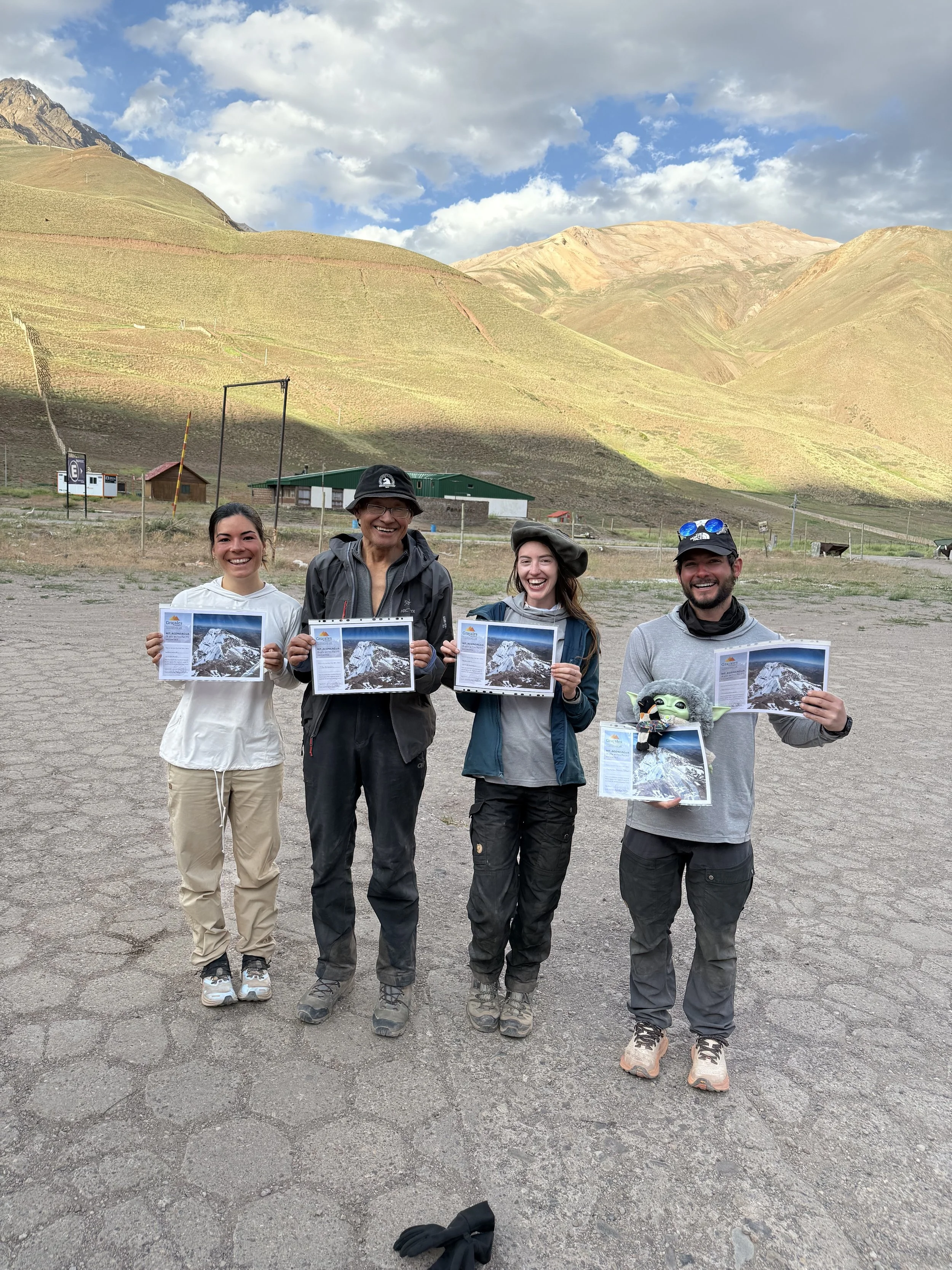 Four people standing on a dirt ground in front of a hilly landscape, each holding a paper with a mountain photo. They are smiling and appear to be on a foggy mountain hike.