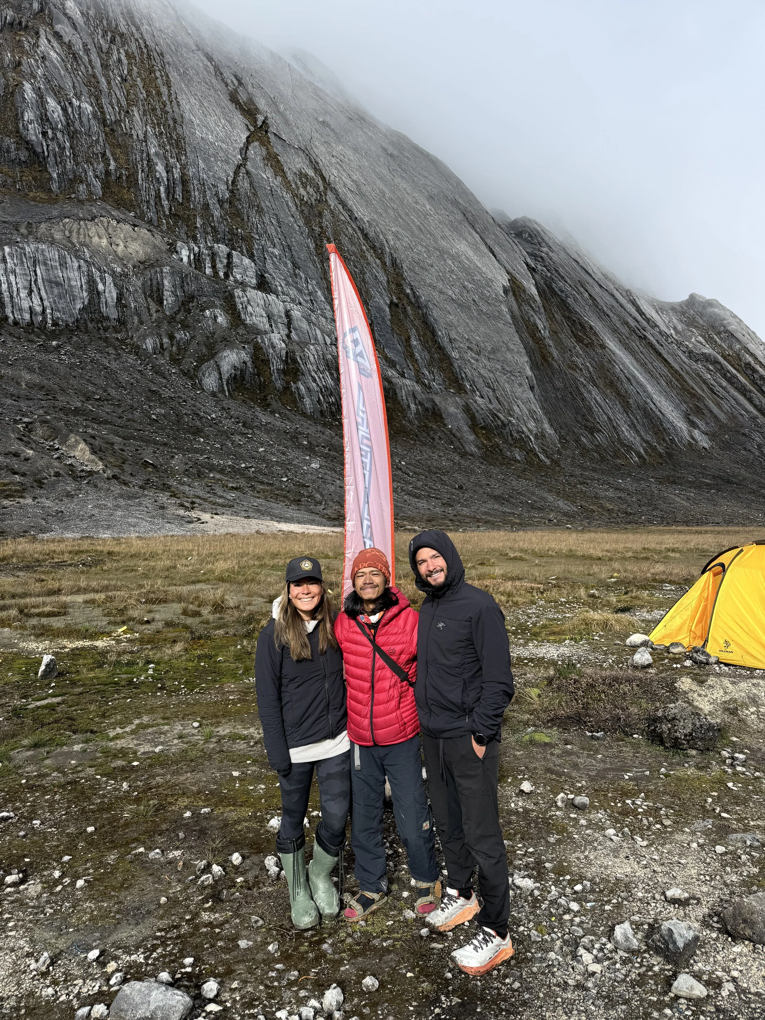 Three people dressed in outdoor gear standing on rocky ground in front of a mountain with fog, with a yellow tent nearby and a tall pink flag behind them.