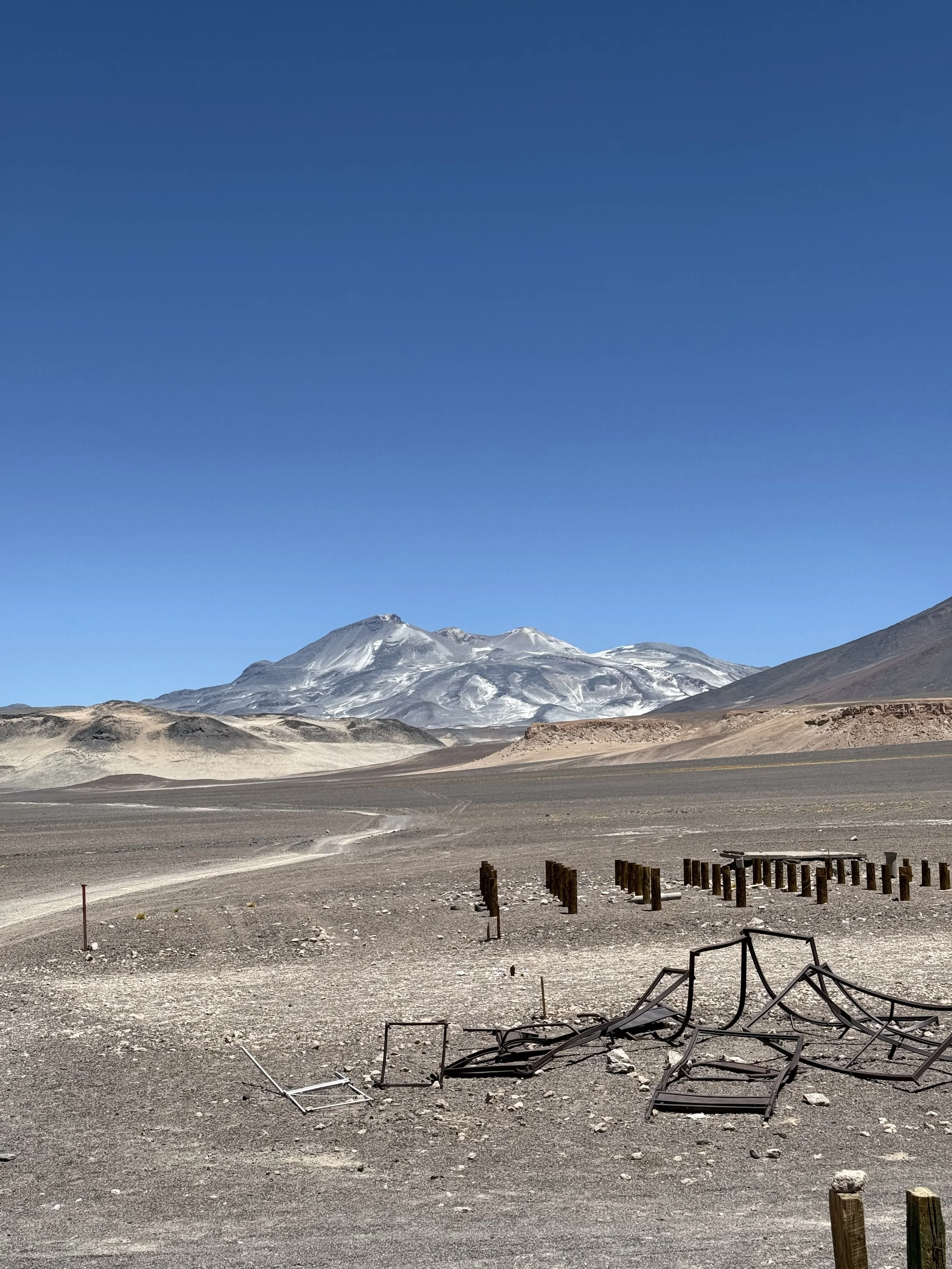 A barren desert landscape with scattered debris, wooden posts, and a snow-capped mountain in the background under a clear blue sky.