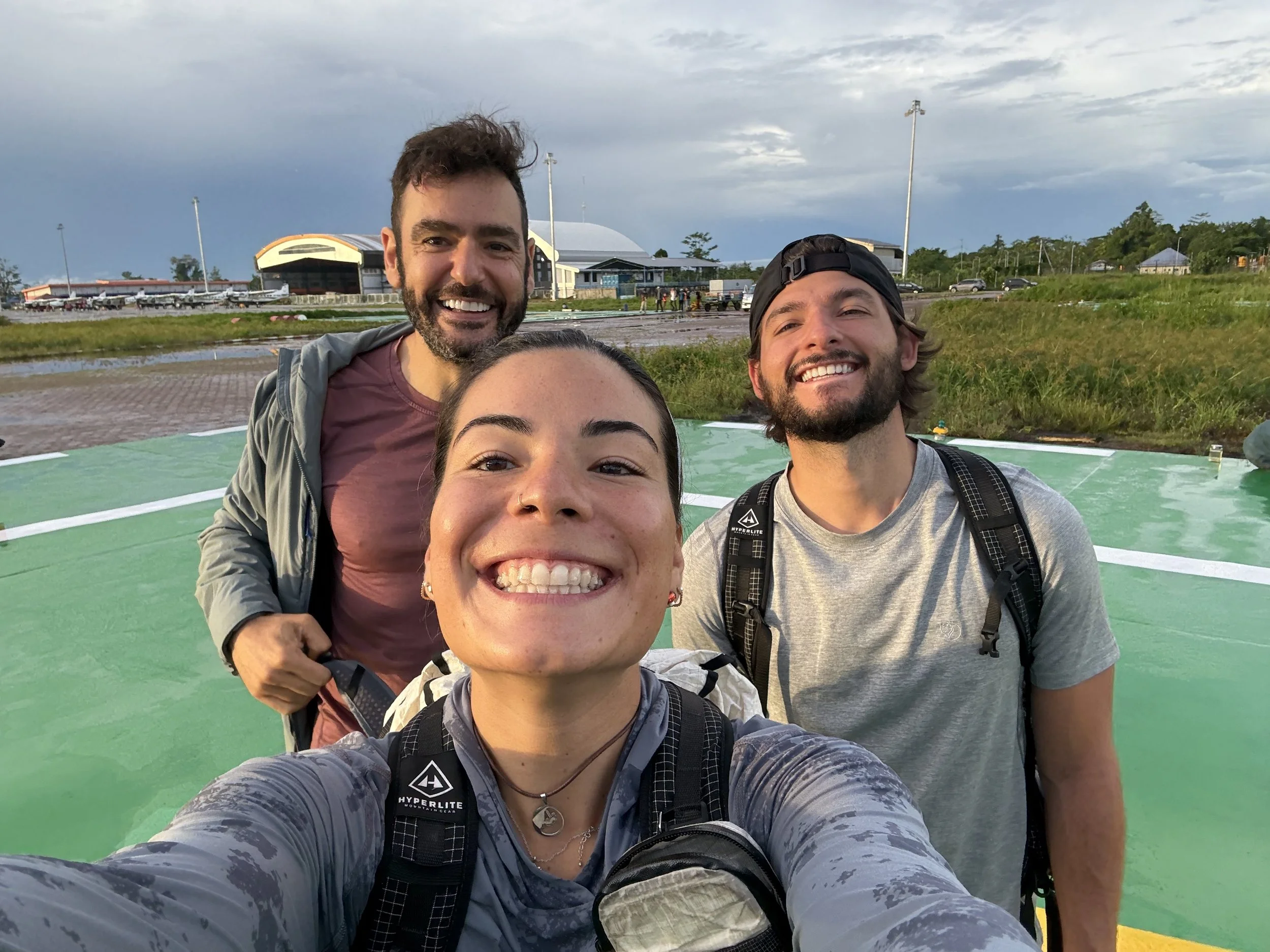 Three smiling young adults taking a selfie outdoors on a green helipad with a cloudy sky and some buildings in the background.