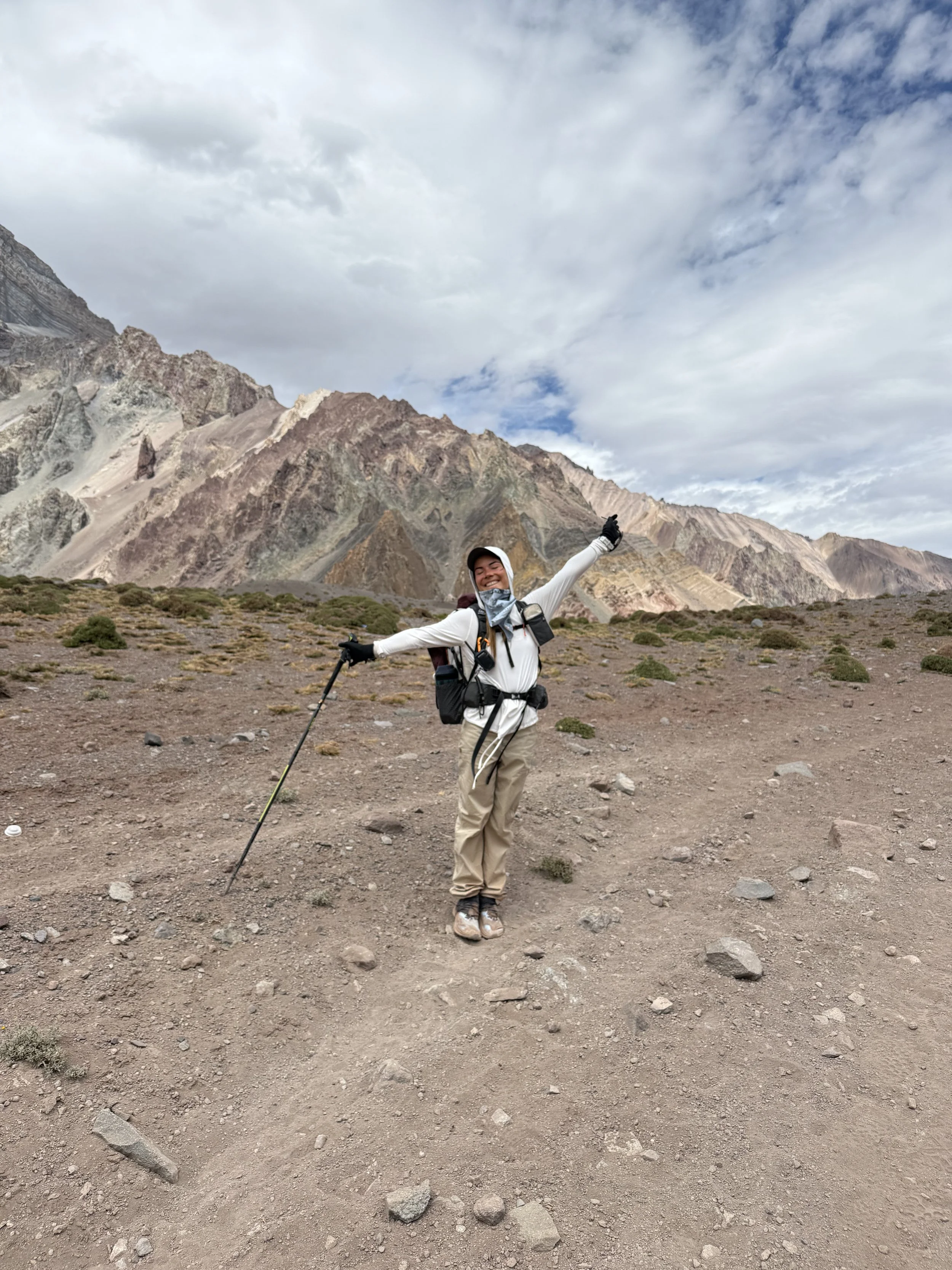 A person standing in a rocky mountain terrain, smiling with arms outstretched, holding a trekking pole, surrounded by mountains and a partly cloudy sky.