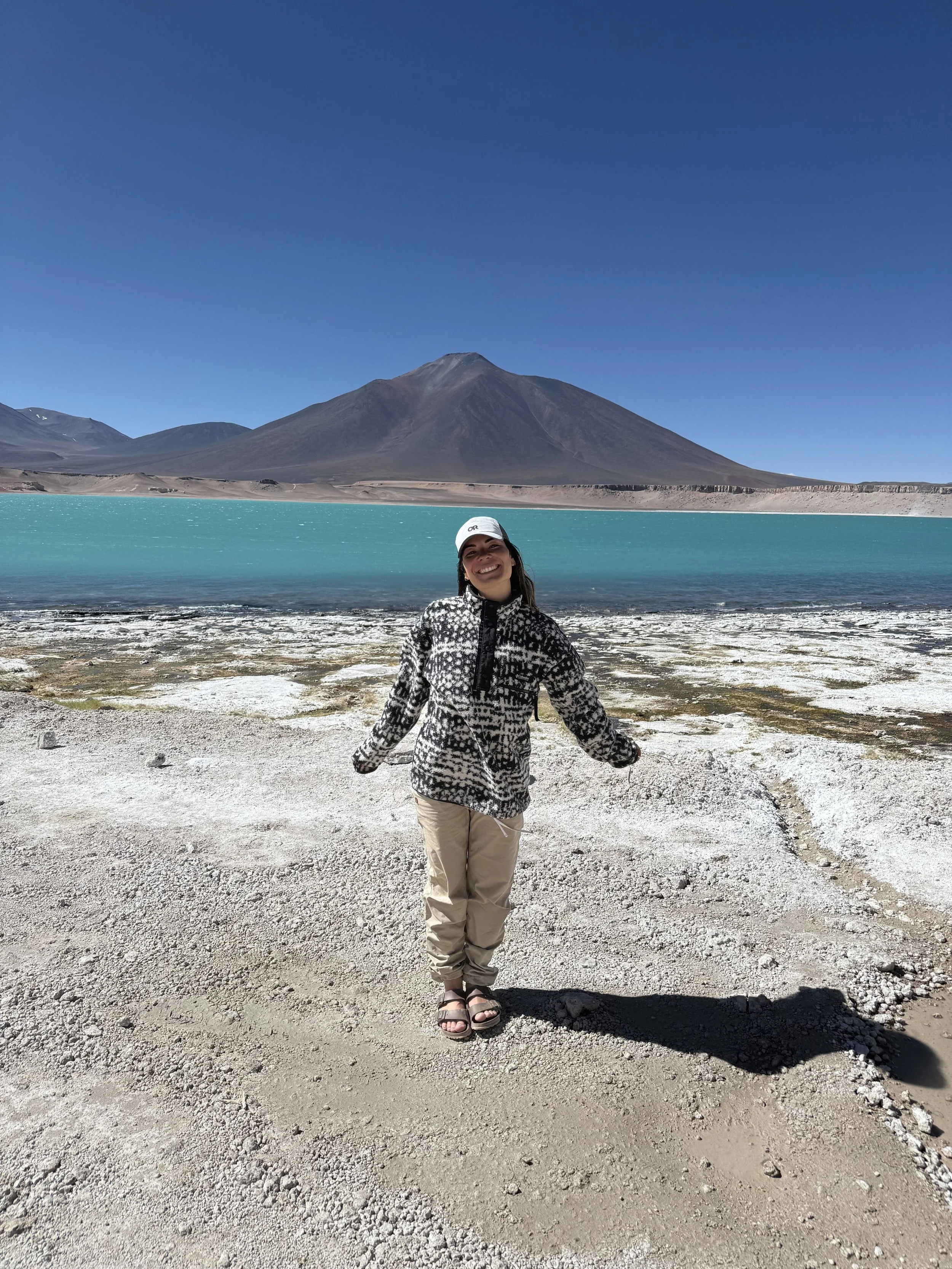 A woman standing on a rocky shore with a mountain and turquoise lake in the background under a clear blue sky.