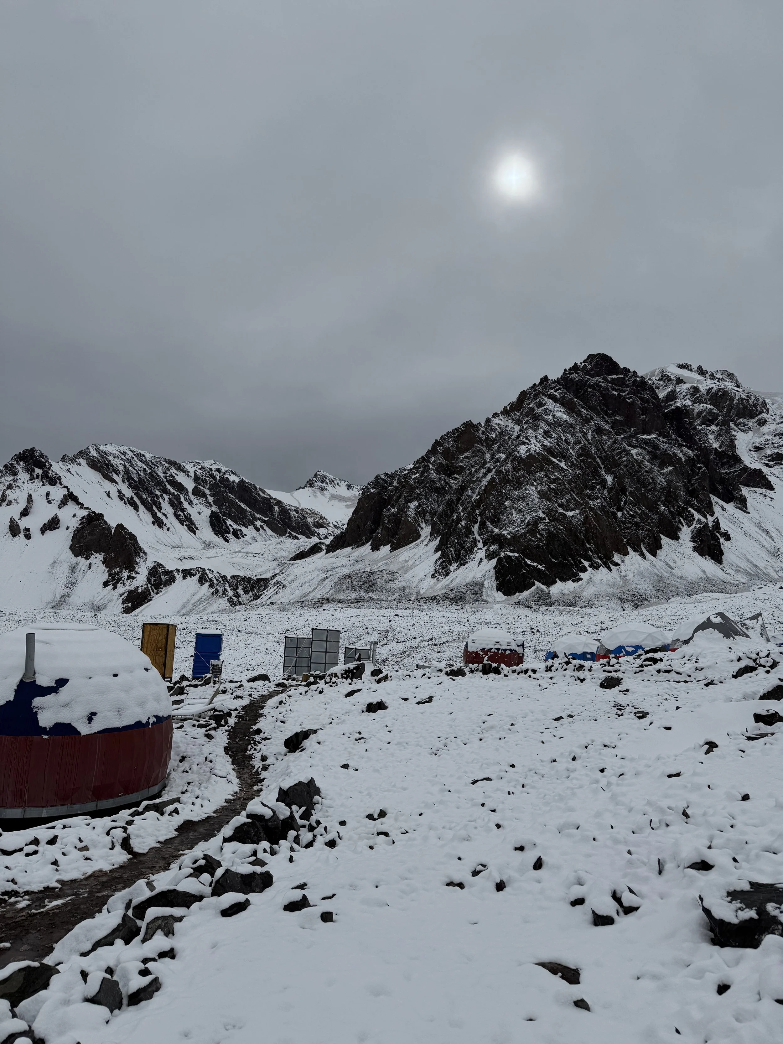 Snow-covered mountainous landscape with small structures and tents in the foreground under gray overcast sky.