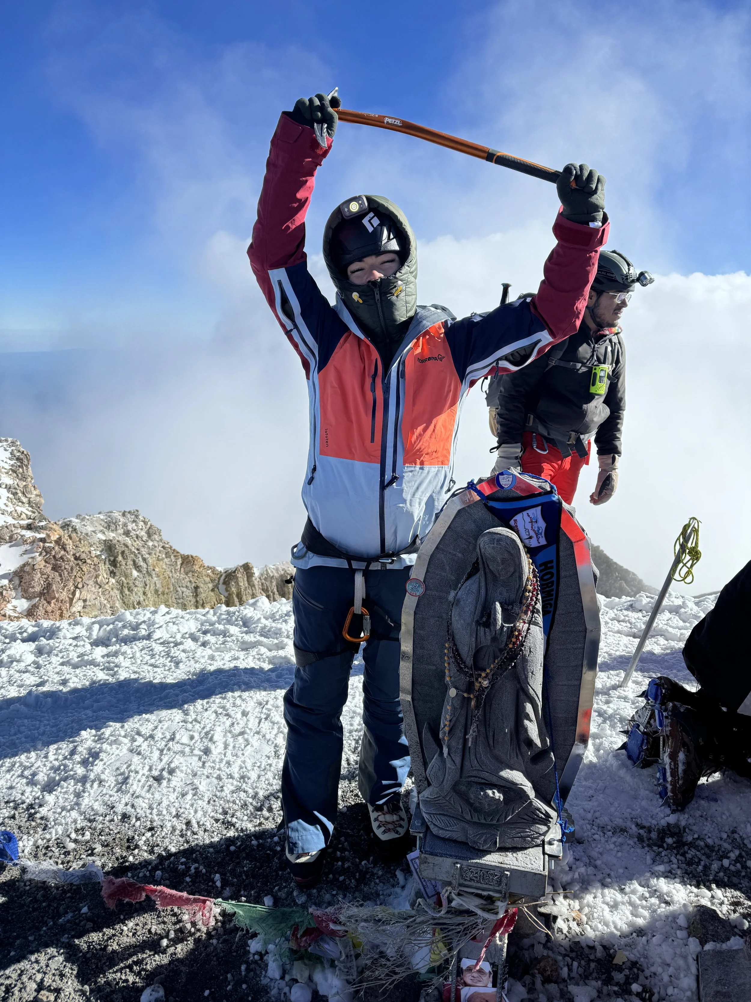 Mountaineer celebrating on snow-covered mountain summit with trekking pole raised, wearing helmet, face covering, colorful jacket, and climbing gear, with clouds in the background.