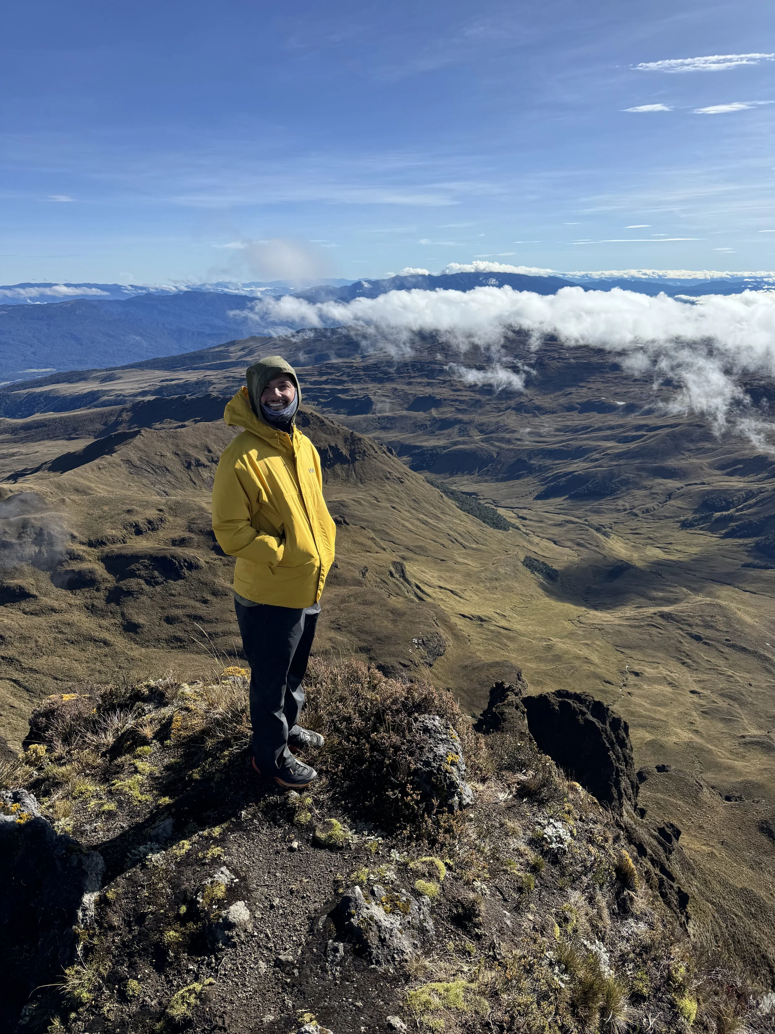 A person wearing a yellow jacket and gray pants standing on a mountain summit with a scenic view of rolling hills and clouds in the background.