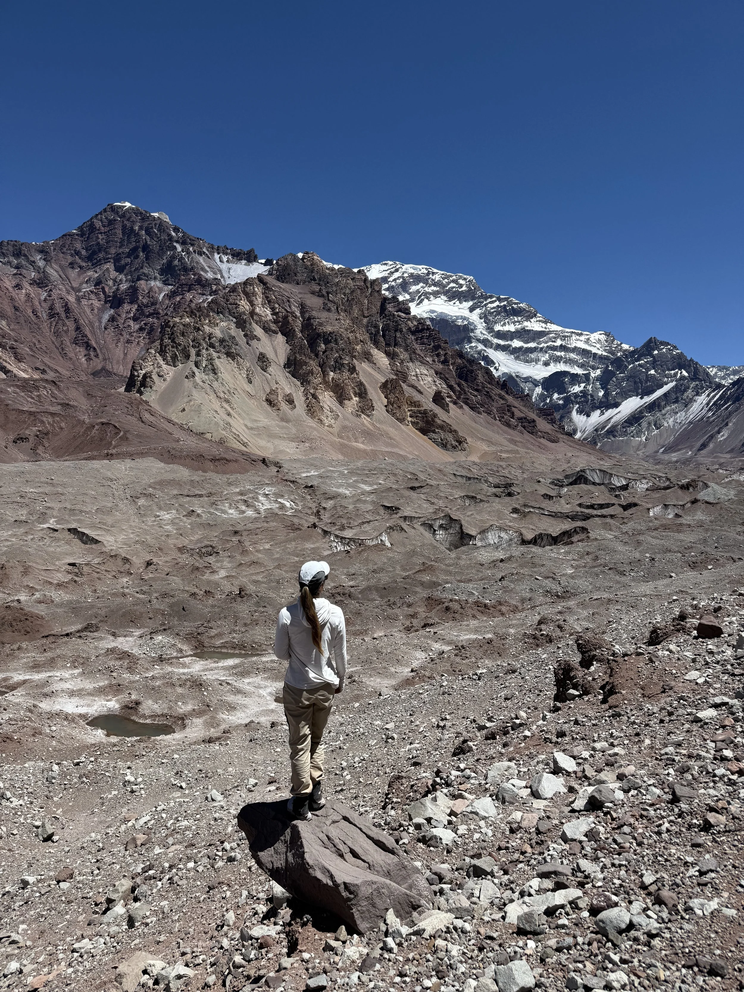 A person in white clothing and a hat standing on a rock in a barren, rocky landscape with tall mountains and snow-covered peaks in the background.