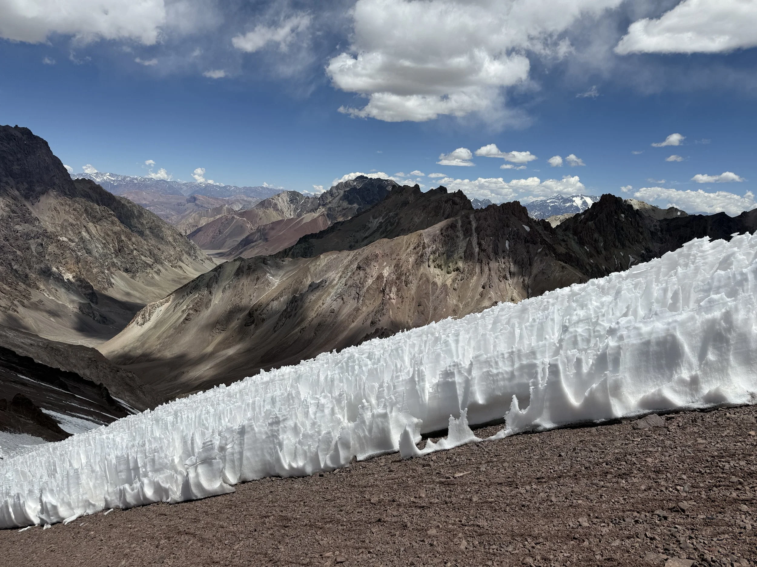 Snowy mountain landscape with a large snowbank in the foreground and rugged mountain peaks in the background under a partly cloudy sky.