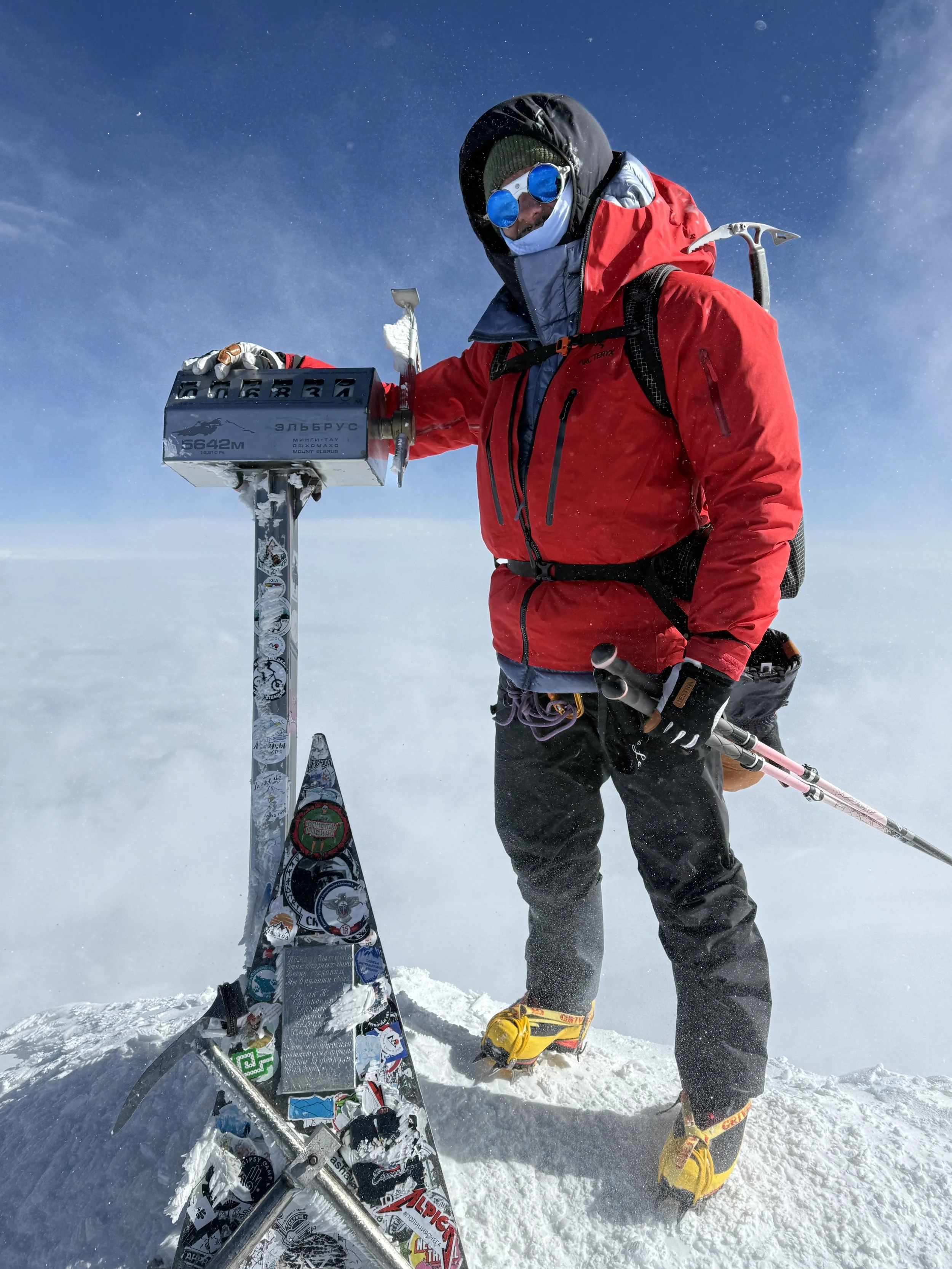 Mountaineer in red jacket with sunglasses and backpack on snowy summit, standing next to a marker with stickers, holding an ice axe and ski poles, with a cloudy sky in the background.