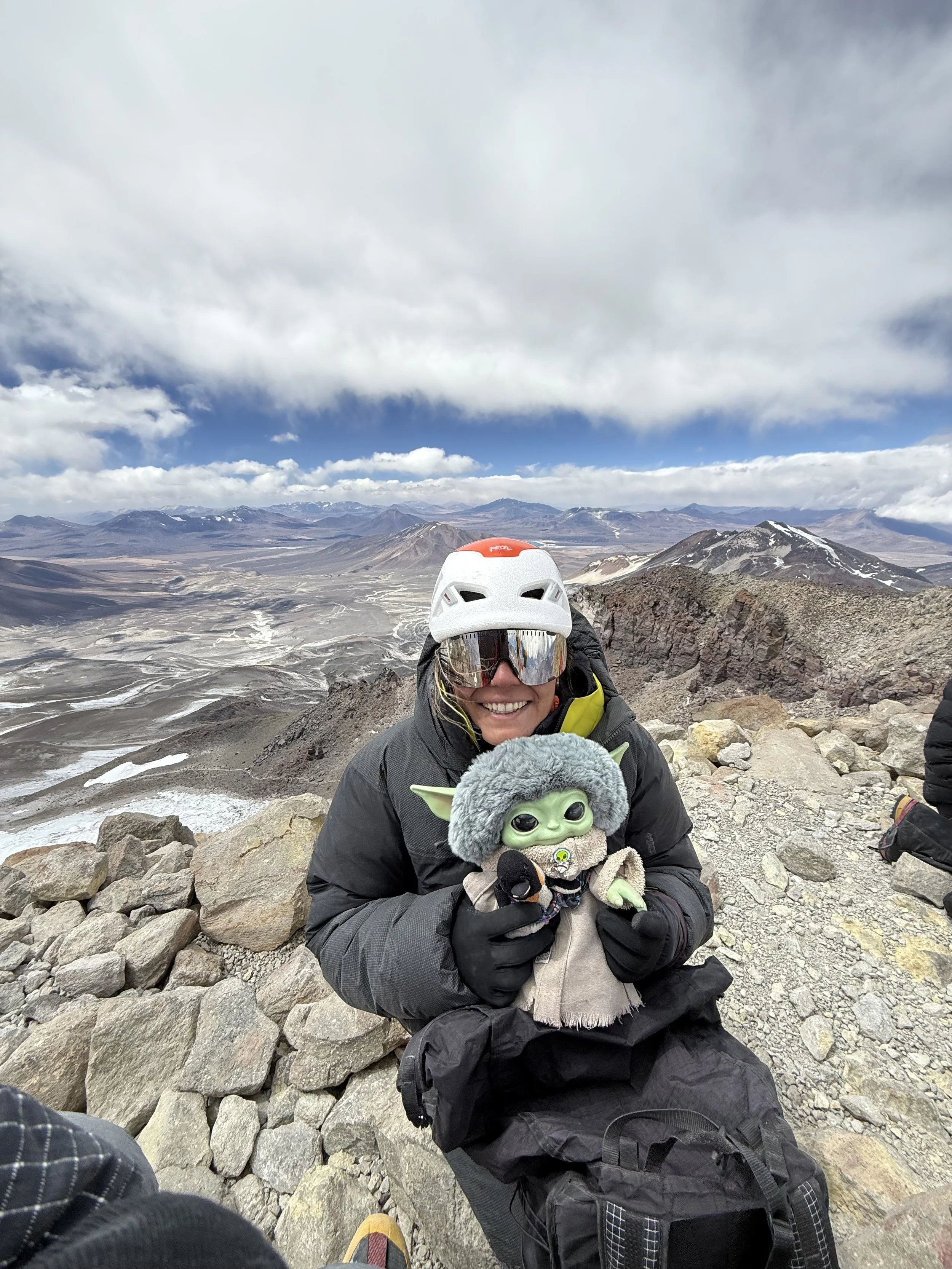 Person in outdoor gear, wearing a helmet and sunglasses, holding a Baby Yoda plush toy, on a rocky mountaintop with a volcanic landscape in the background.
