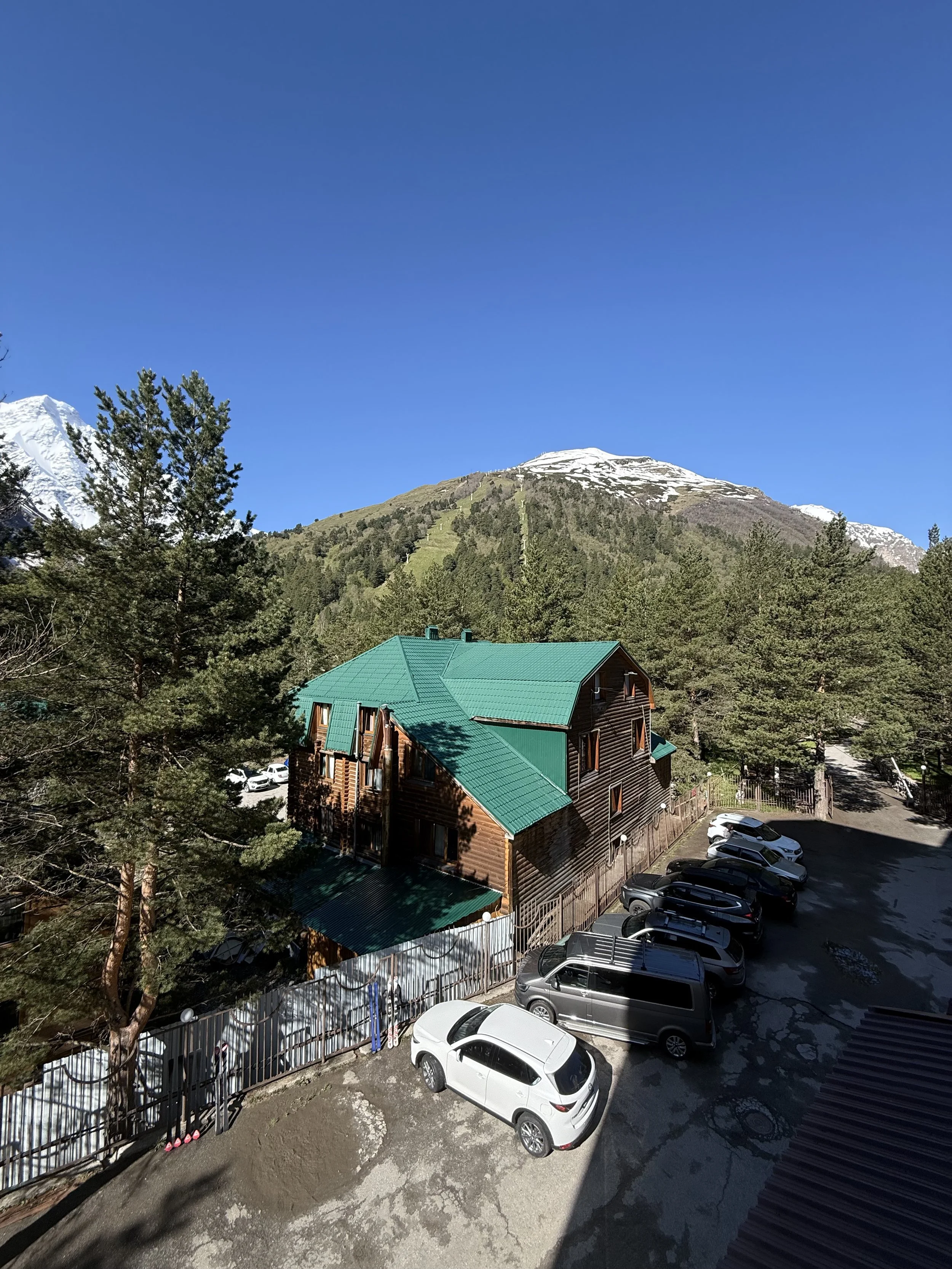 A wooden building with a green metal roof surrounded by pine trees, parked cars, and snow-capped mountains in the background under a clear blue sky.