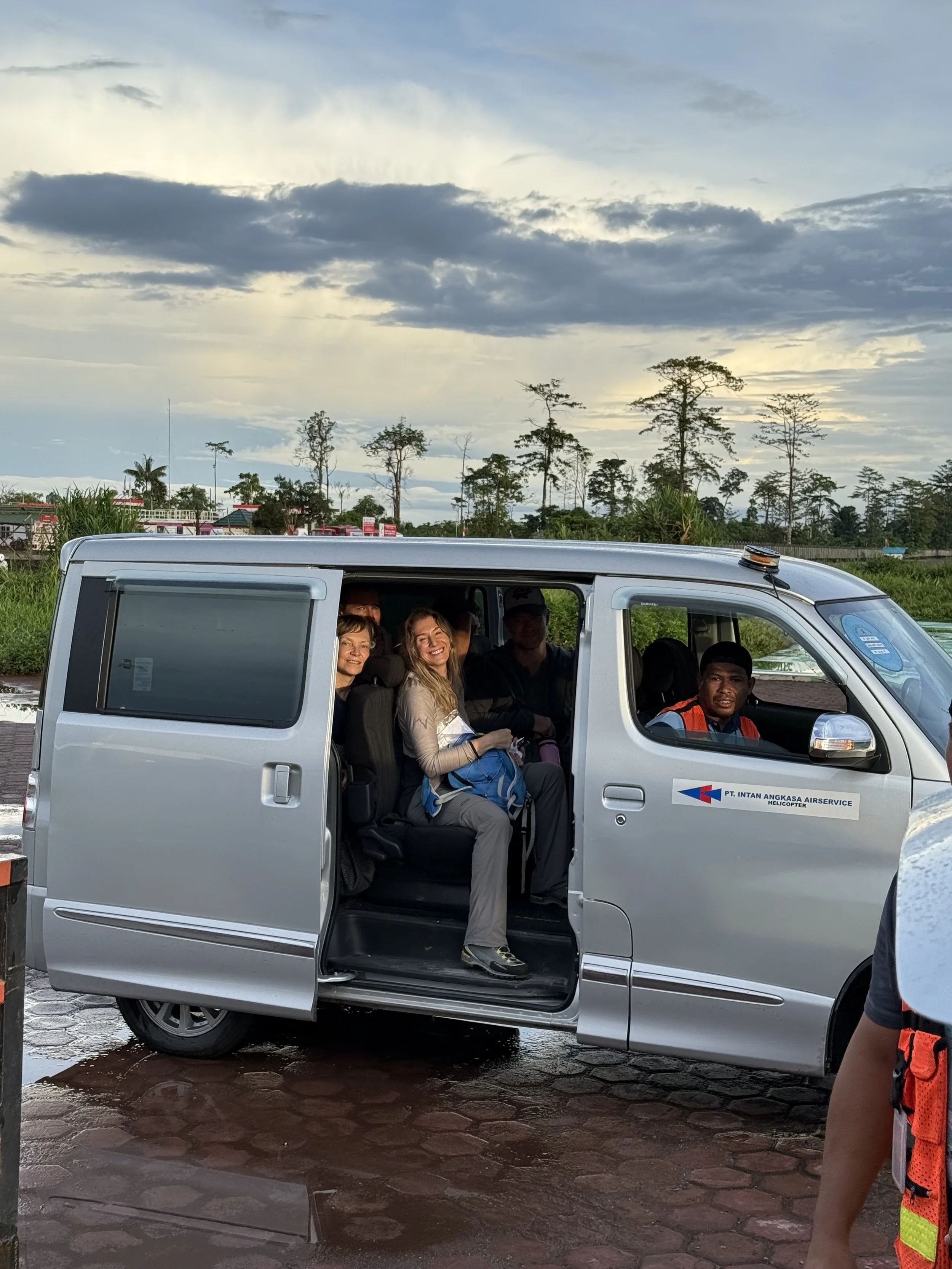 People sitting inside a parked gray van at an outdoor location during sunset, with trees and cloudy sky in the background.