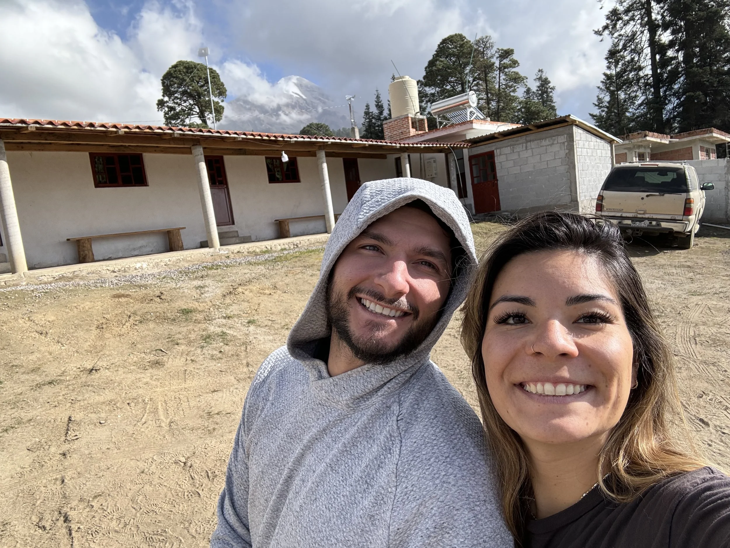 Smiling couple taking a selfie outdoors with a mountain in the background, a single-story building, a parked vehicle, trees, and partly cloudy sky.