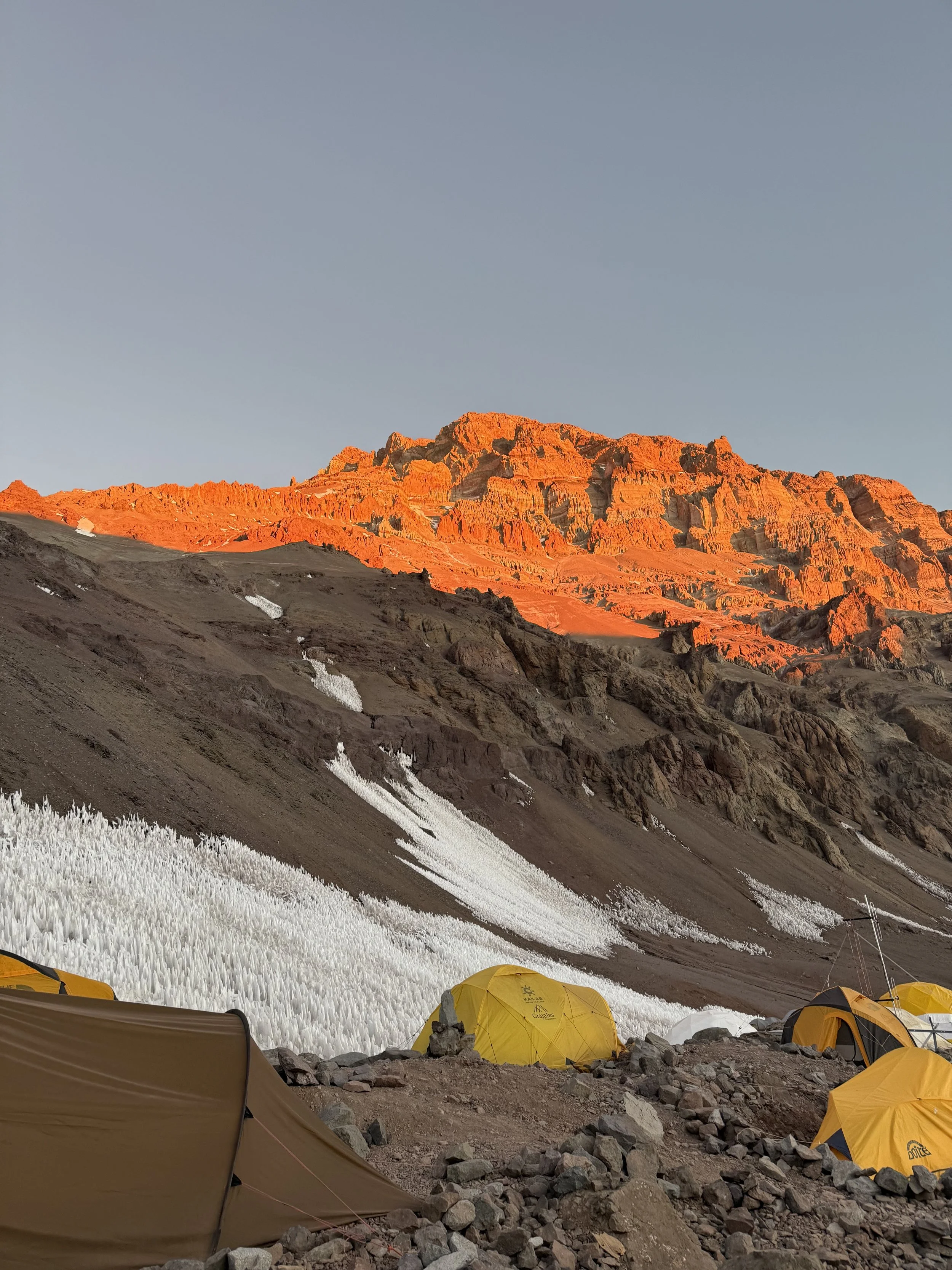 A mountain landscape with orange-lit peaks in the background and a campsite with yellow tents in the foreground.