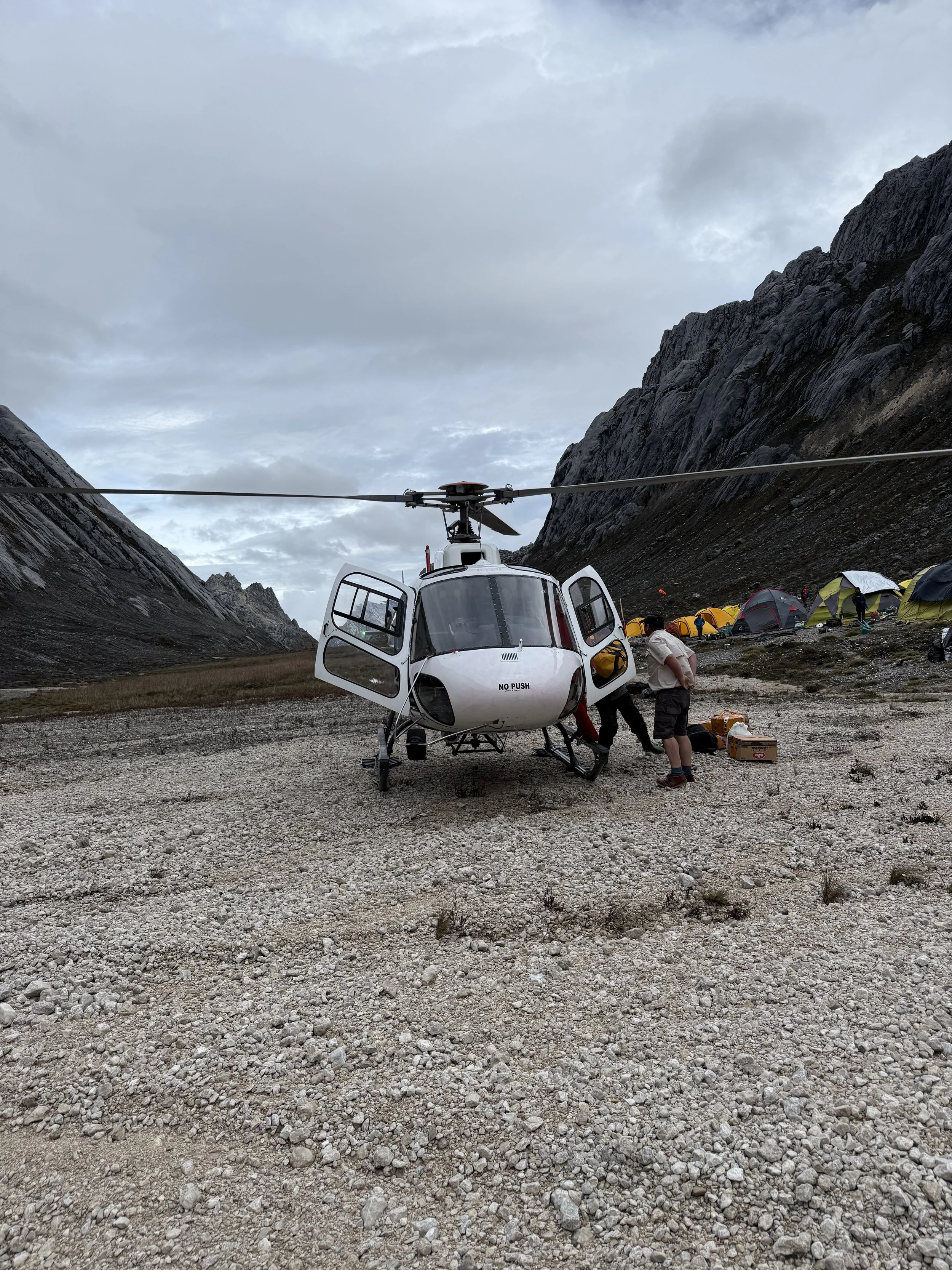 A white helicopter with open doors parked on rocky terrain surrounded by tents and mountains in a remote outdoor setting.