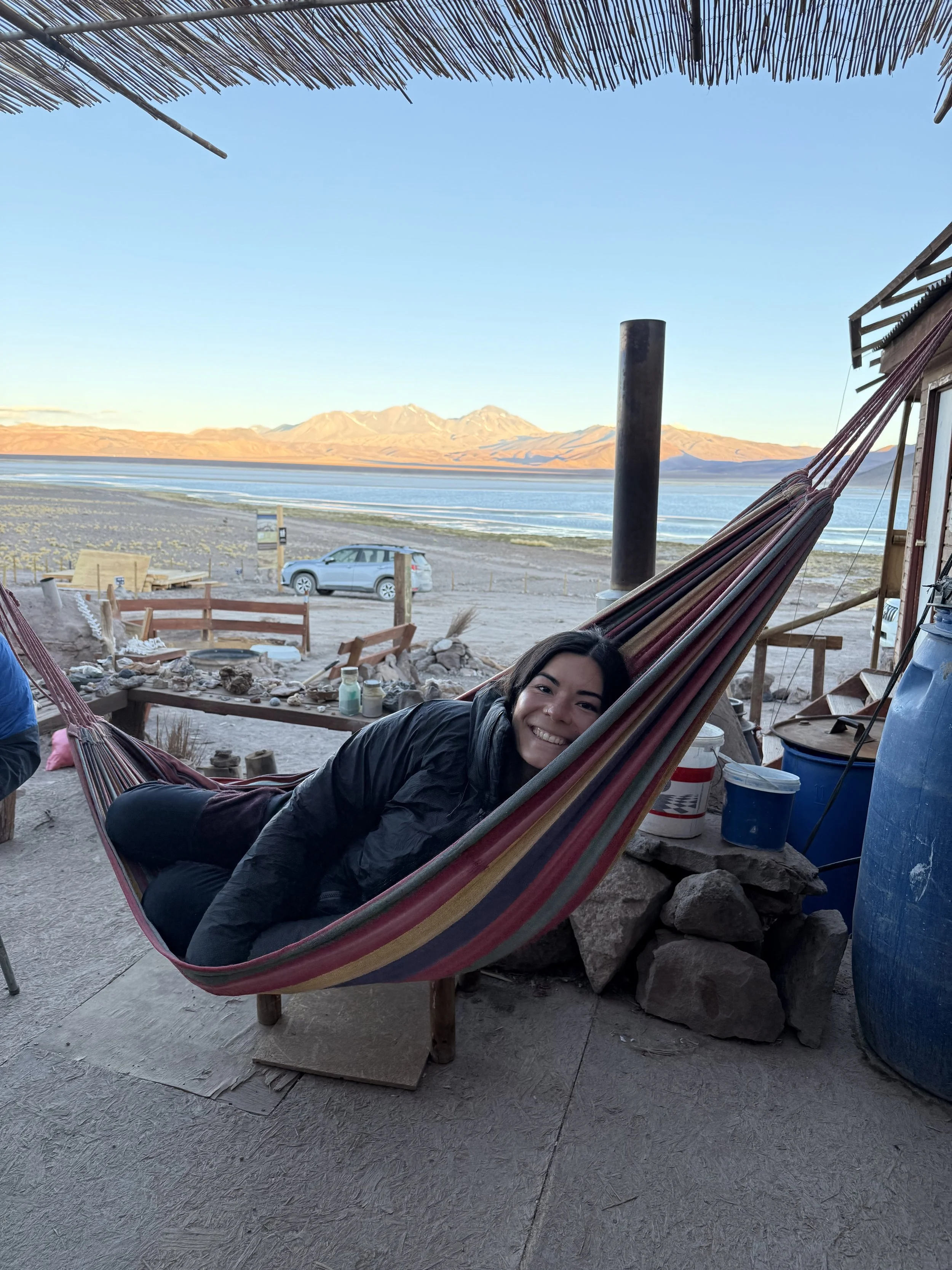 Woman lying in a striped hammock outdoors with mountains, water, and a car in the background during daytime.