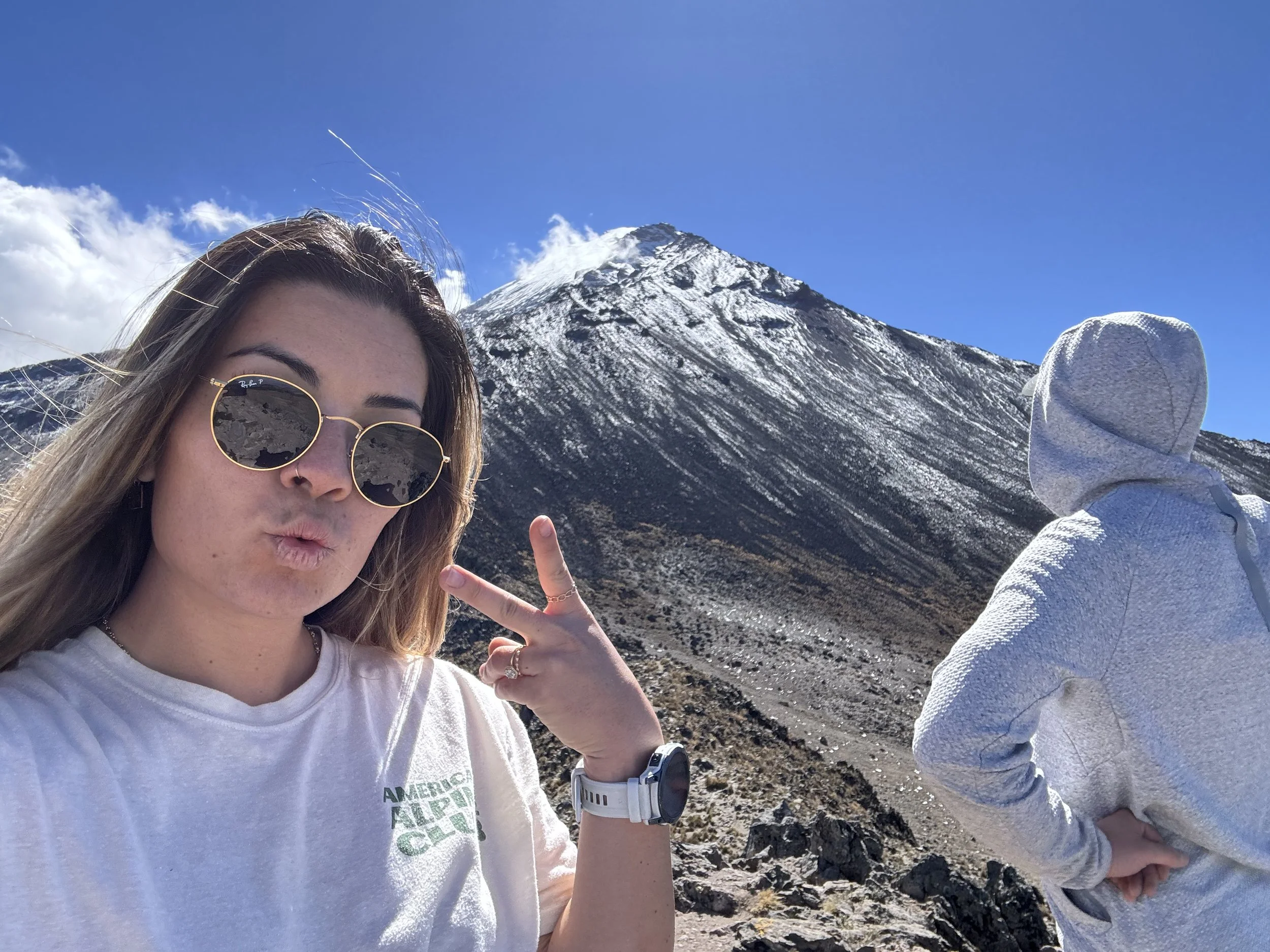 Young woman in sunglasses making a peace sign in front of a snow-capped mountain, with another person in a gray hoodie looking at the mountain.